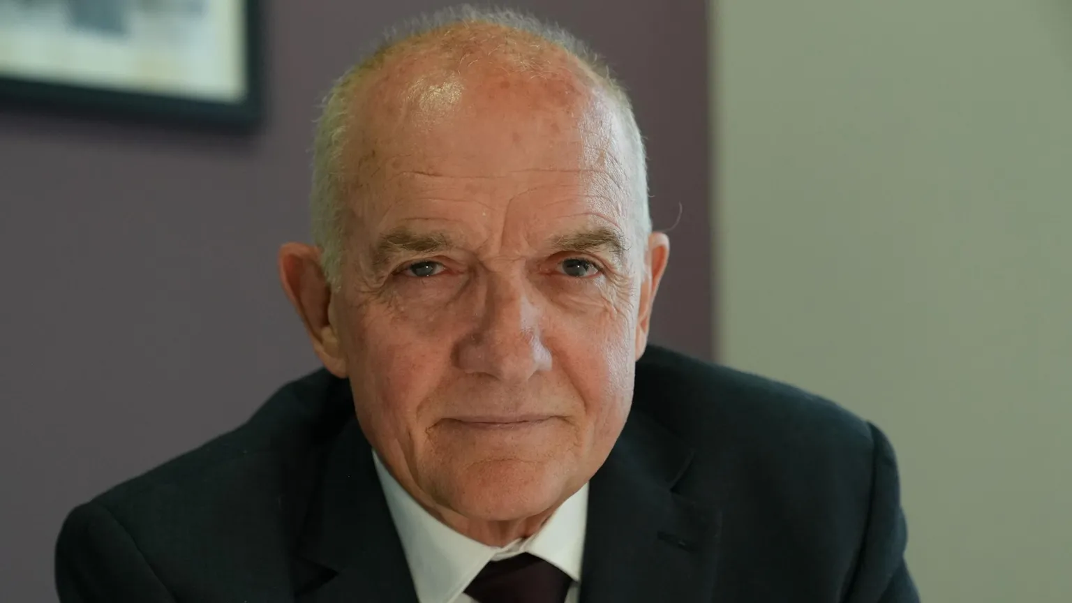 Colin Anderson looking directly at the camera. He has grey hair and is wearing a black suit, white shirt and black tie.