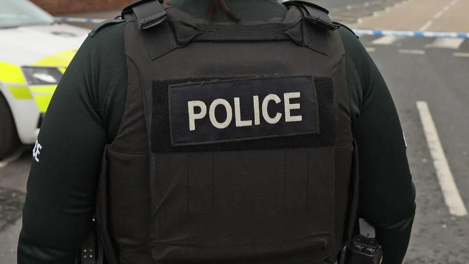  A close-up image of a police officer's black protection vest as they stand near a cordoned off street.