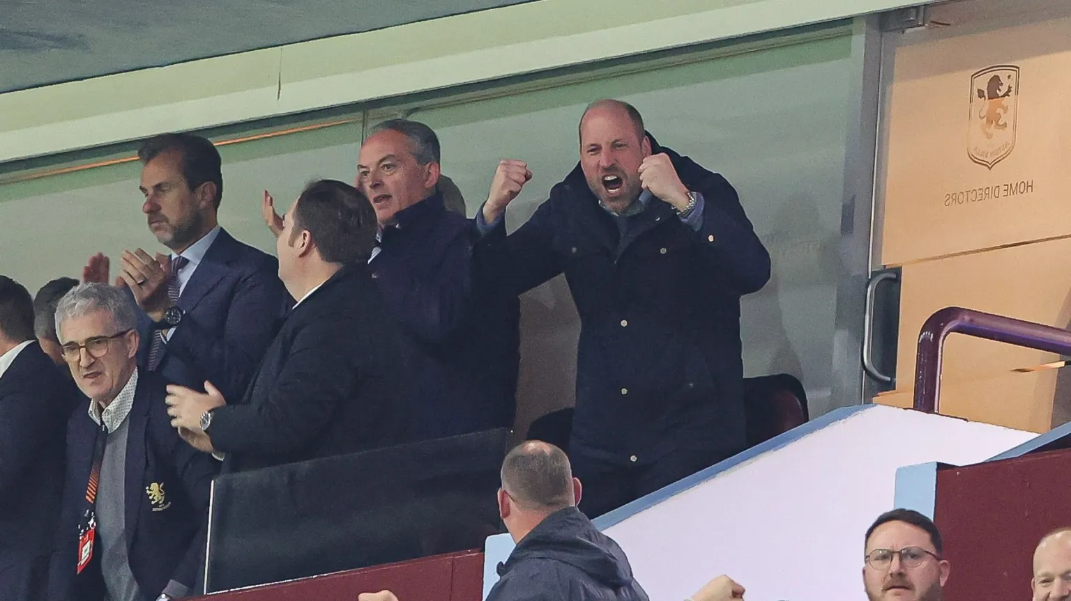  Prince William is standing with his fists in the air in celebration, with his face mid-cheer. There is a group of men to his right that all appear to be clapping