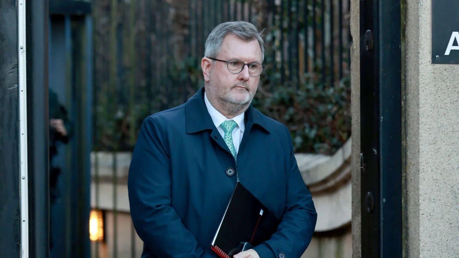 Sir Jeffrey Donaldson, a man with grey hair and dark glasses, wearing a blue jacket, white shirt and green tie, holding a black and red book.