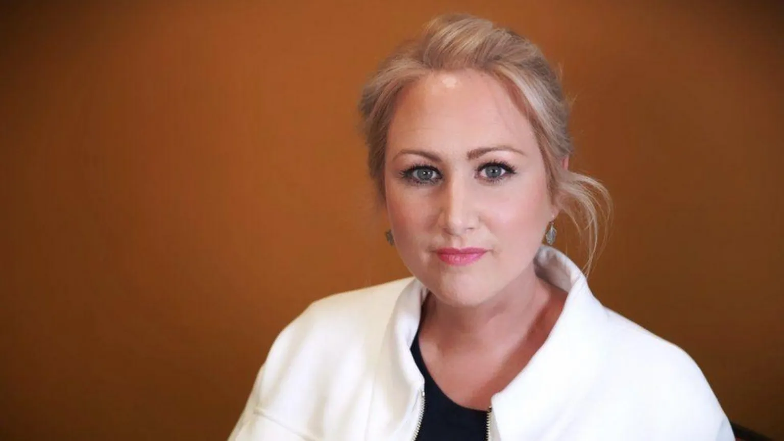 S4C Llinos Griffin-Williams standing in front of an orange background. She is looking at the camera. She has her hair tied back and is wearing a white jacket.
