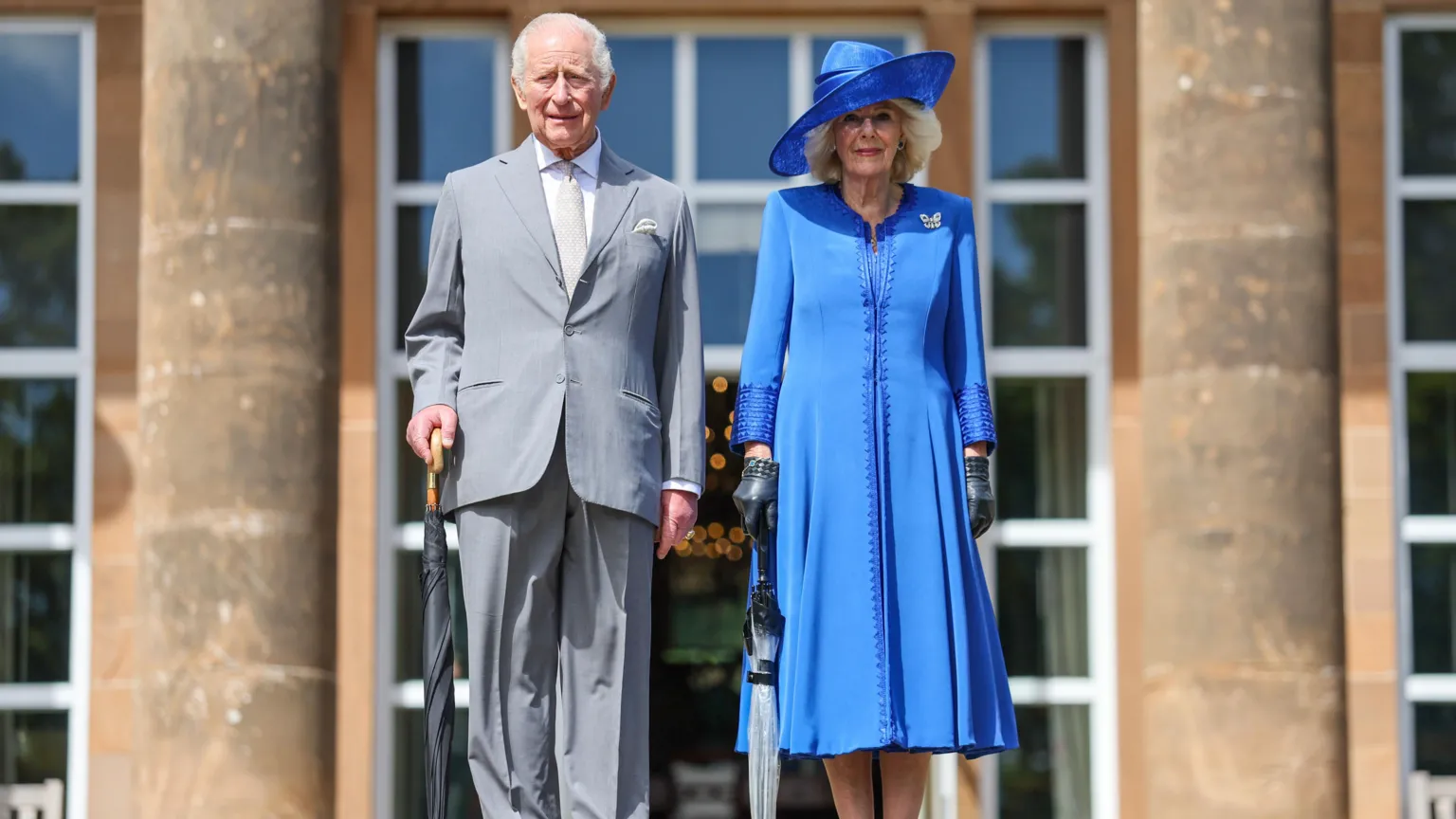 The King stands on the right of Camilla. He wears a grey suit and trousers, with a white shirt and grey tie. He has short grey hair. Camilla wears a knee-length blue dress with black gloves and a round blue hat that shades her face. Both have umbrellas in their right hands.