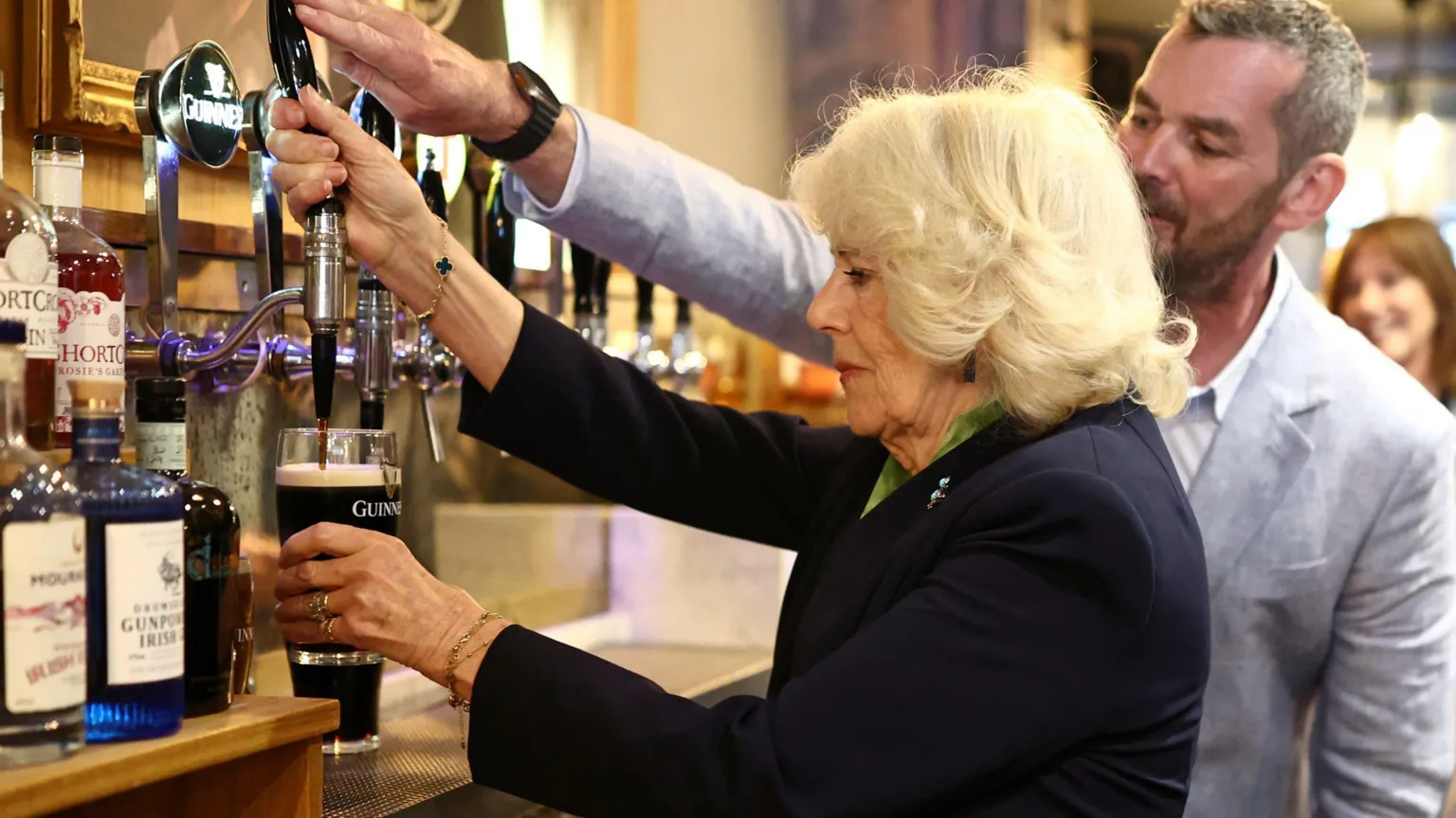 Queen Camilla wears a navy blazer and green shirt as she holds a partially full glass of black liquid, with a white head, in her left hand. She holds a black tap with her right hand. A man with short grey hair and a short brown beard helps her, his right hand is on the top of the tap. He wears a grey linen suit and a white shirt.