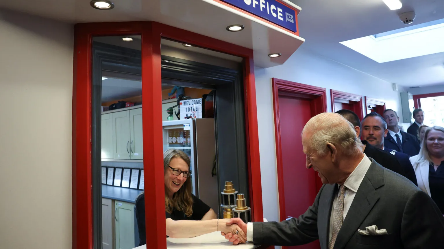 The King shakes hands with a woman with long brown hair, who wears a black t-shirt and glasses. She stands at the box office of a cinema. The king is wearing a grey suit, white shirt and grey tie. Behind the king are half a dozen [people who watch on.