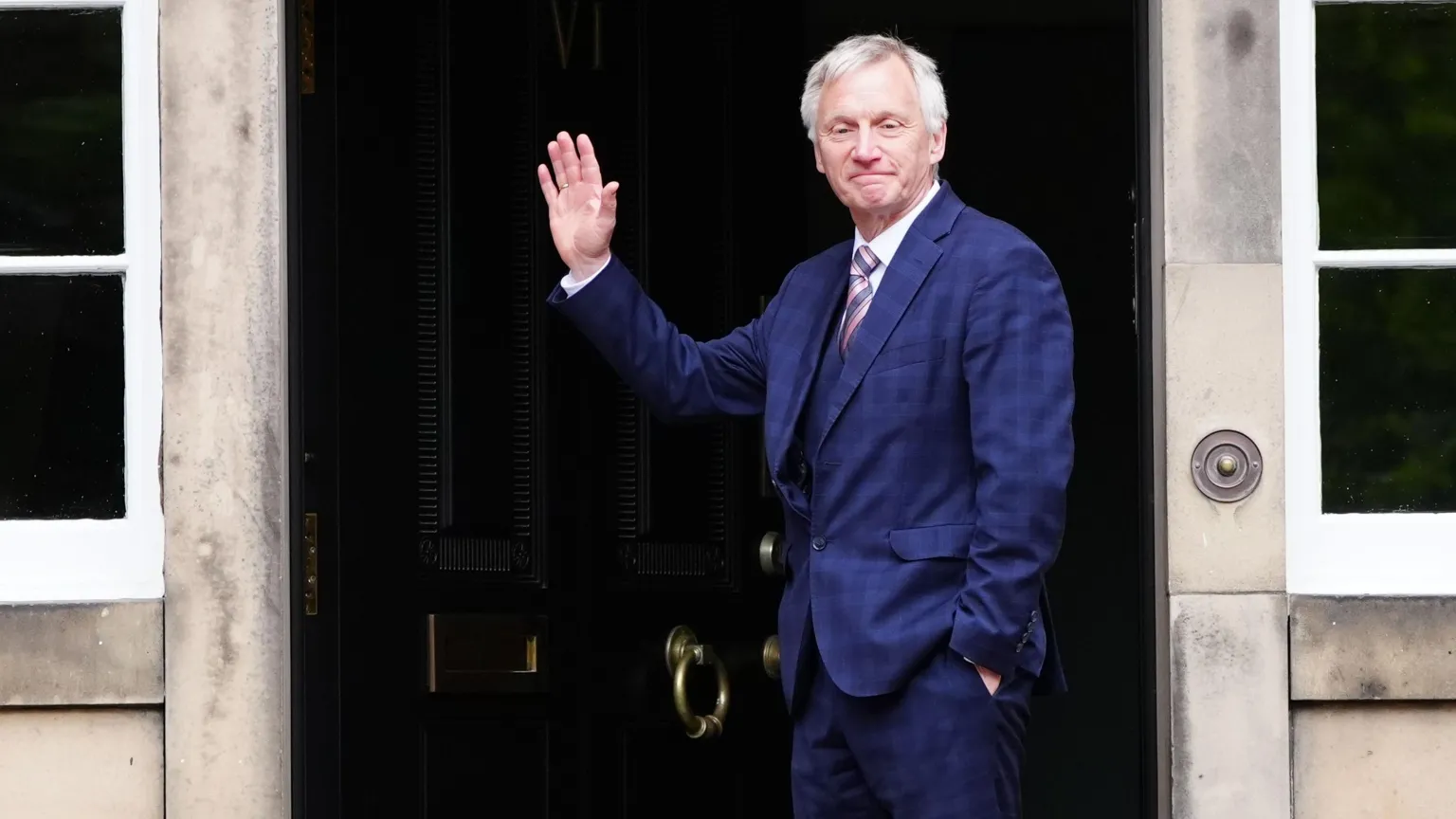  Ivan McKee, wearing a blue checked suit and a pink striped tie waves as he stands in front of a black door.