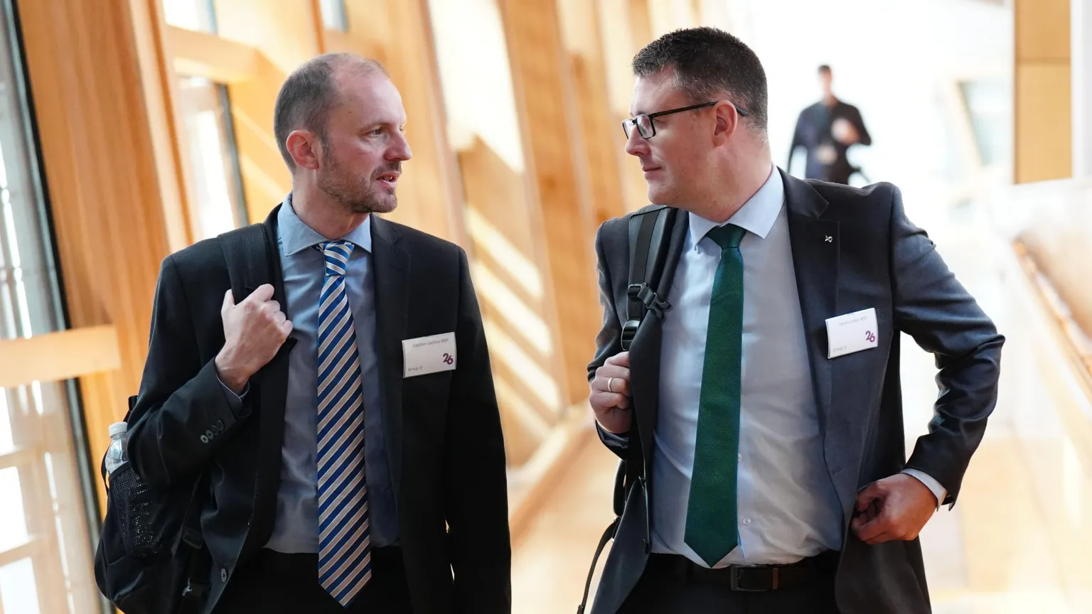  Stephen Gethins wearing a dark suit, blue shirt and striped tie. He is hold a rucksack over his shoulder. He is walking down a corridor in Holyrood next to David Linden, also wearing a suit with a green tie.
