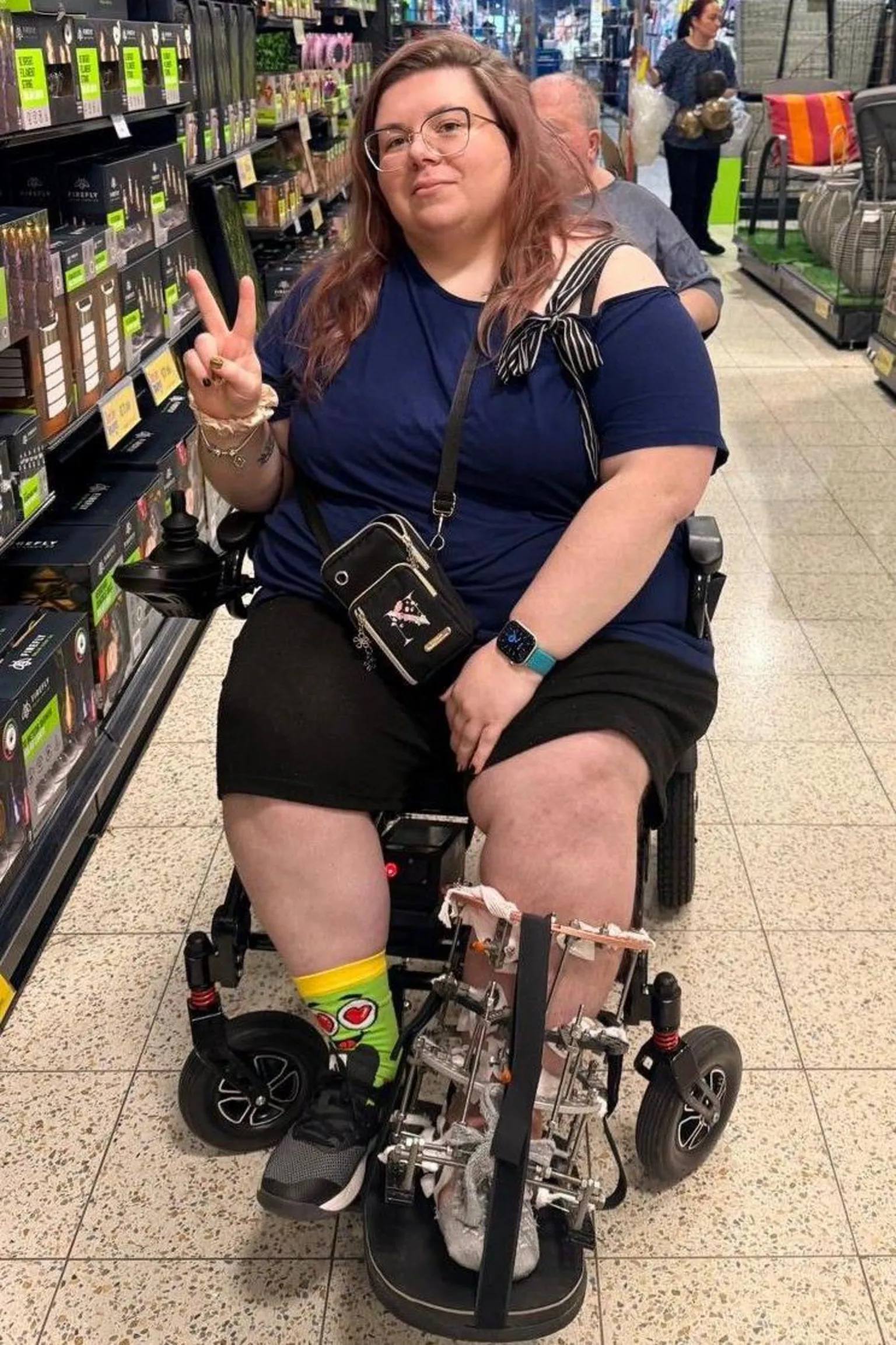 Natalia Sidorska Natalia sits on a wheelchair inside a supermarket aisle. Her left leg is in a metal cage and she makes a peace sign pose. She has long hair, which is dyed a faded pink colour.