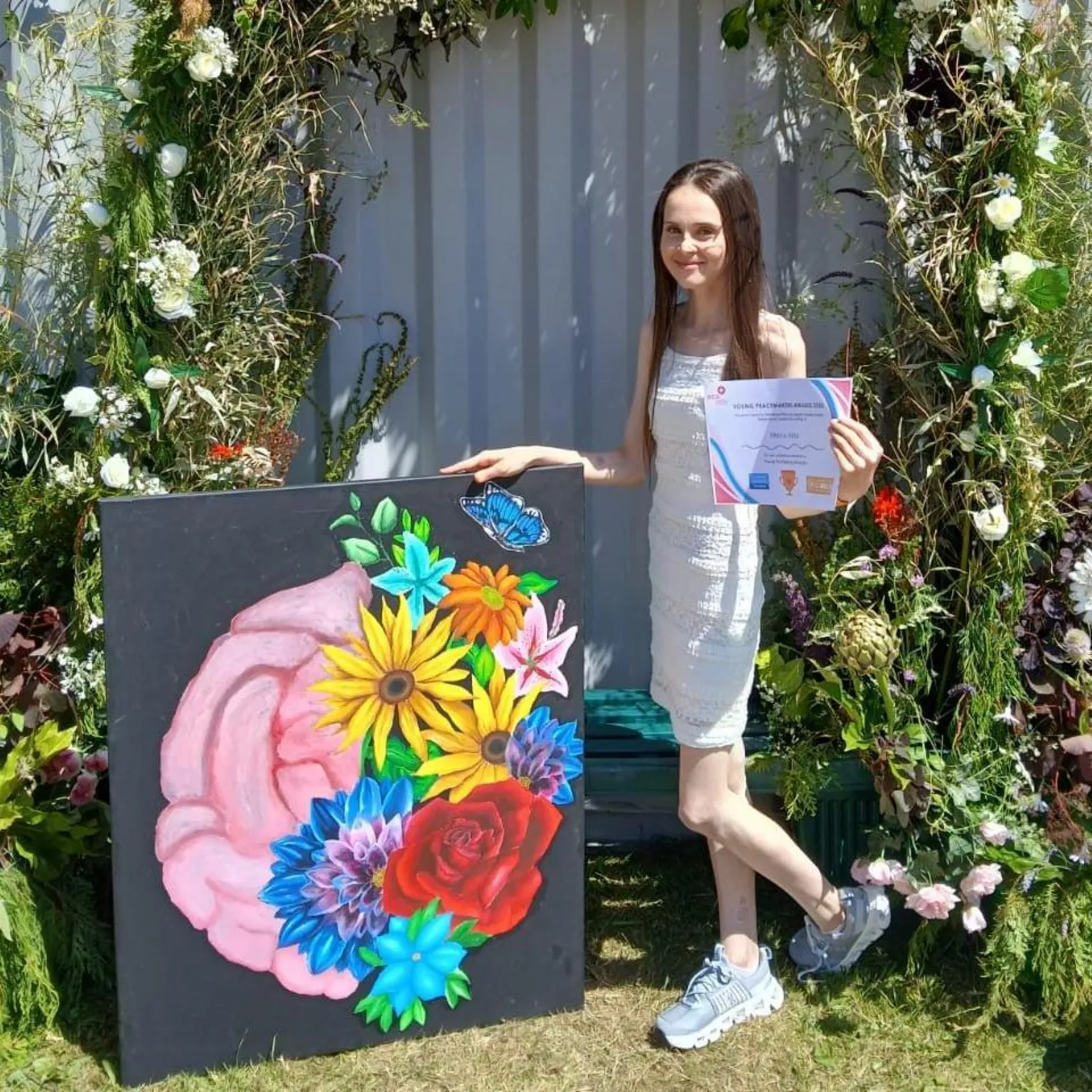 Victoria Glover Freya is wearing a white dress and has long, straight brown hair. Freya is standing next to a colourful piece of artwork. The painting is of part of a brain on the left side and vibrant flowers on the right side. Freya is holding a certificate as this was the artwork that won the prize at the Llangollen Eistedfodd.
