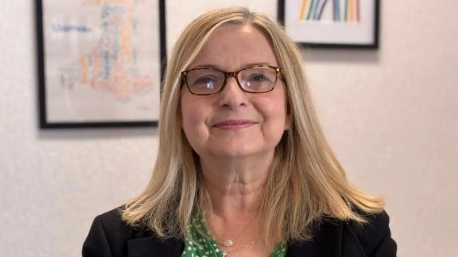 Sam Austin is smiling directly at the camera. She is a white women in her 50's. Sam is wearing rectangular glasses and has shoulder length, blonde hair. She is wearing a green top with white polkadots on it and a black blazer over her shoulders.