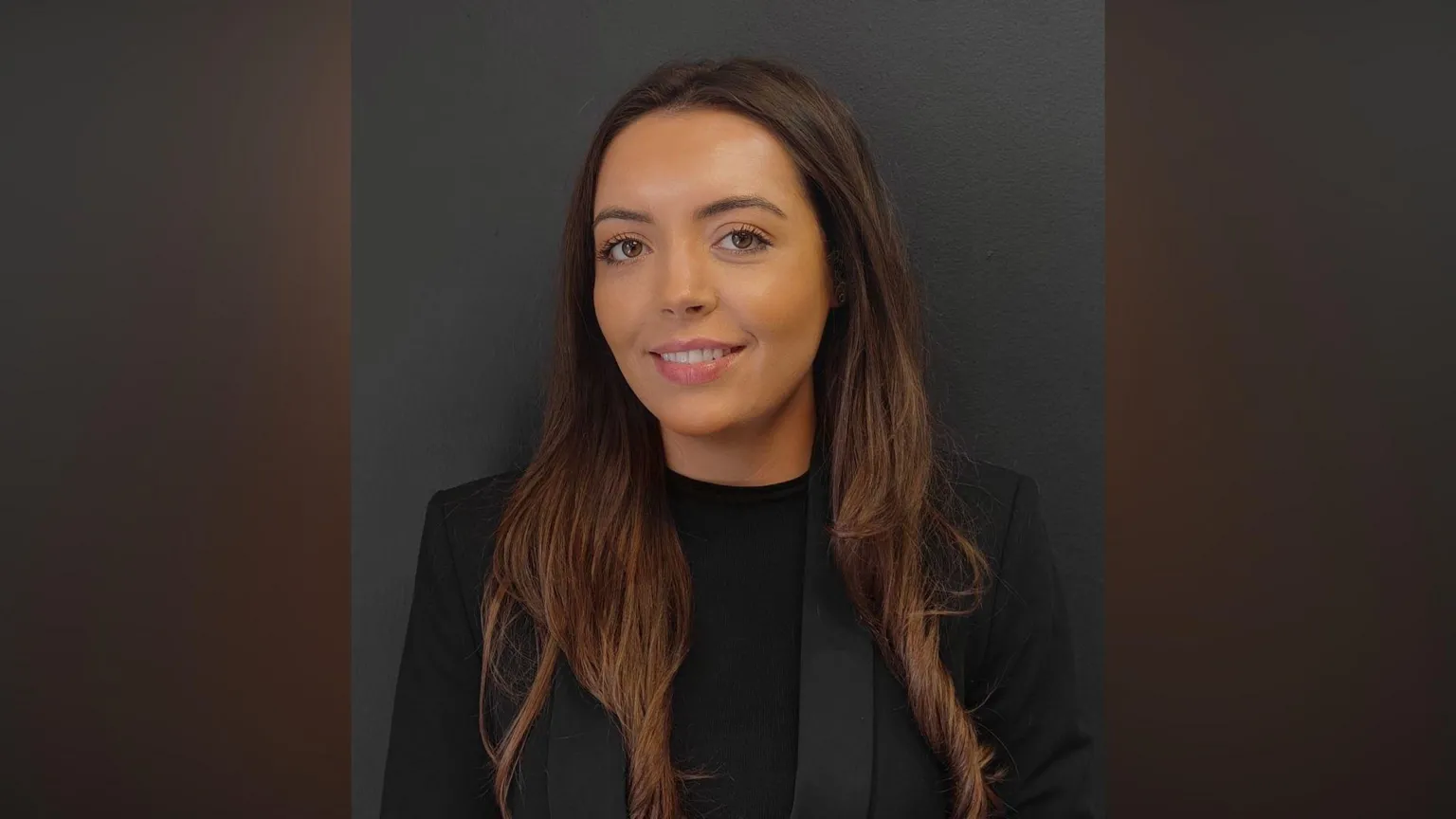 Phoenix Law Victoria smiling at the camera. She has long brown hair and brown eyes. She is wearing a black top under a black blazer and is standing in front of a black wall.