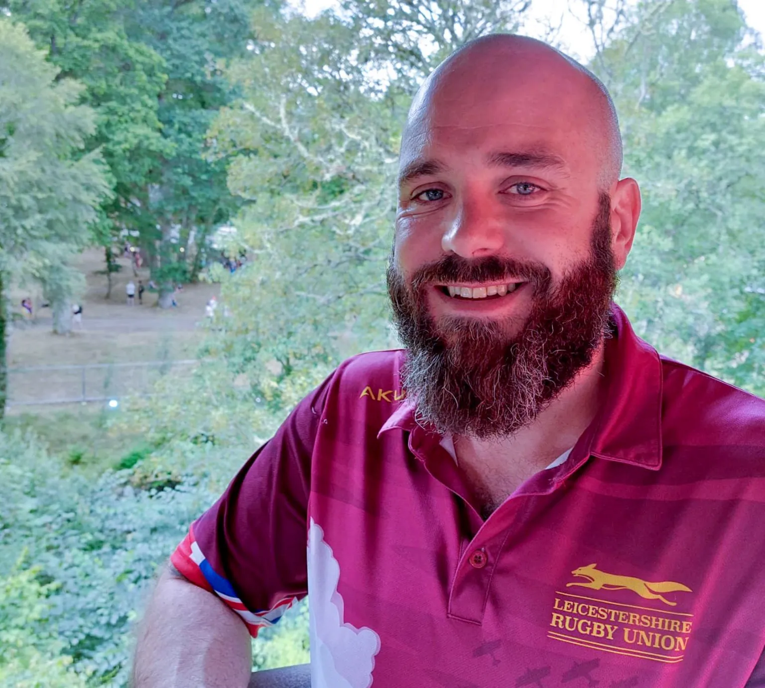 Catriona Ball Lewis is smiling as he stands outside. Trees can be seen in the distance. He is bald and has a long brown and grey beard and moustache. He is wearing a Leicestershire Rugby Union maroon top with yellow logo.