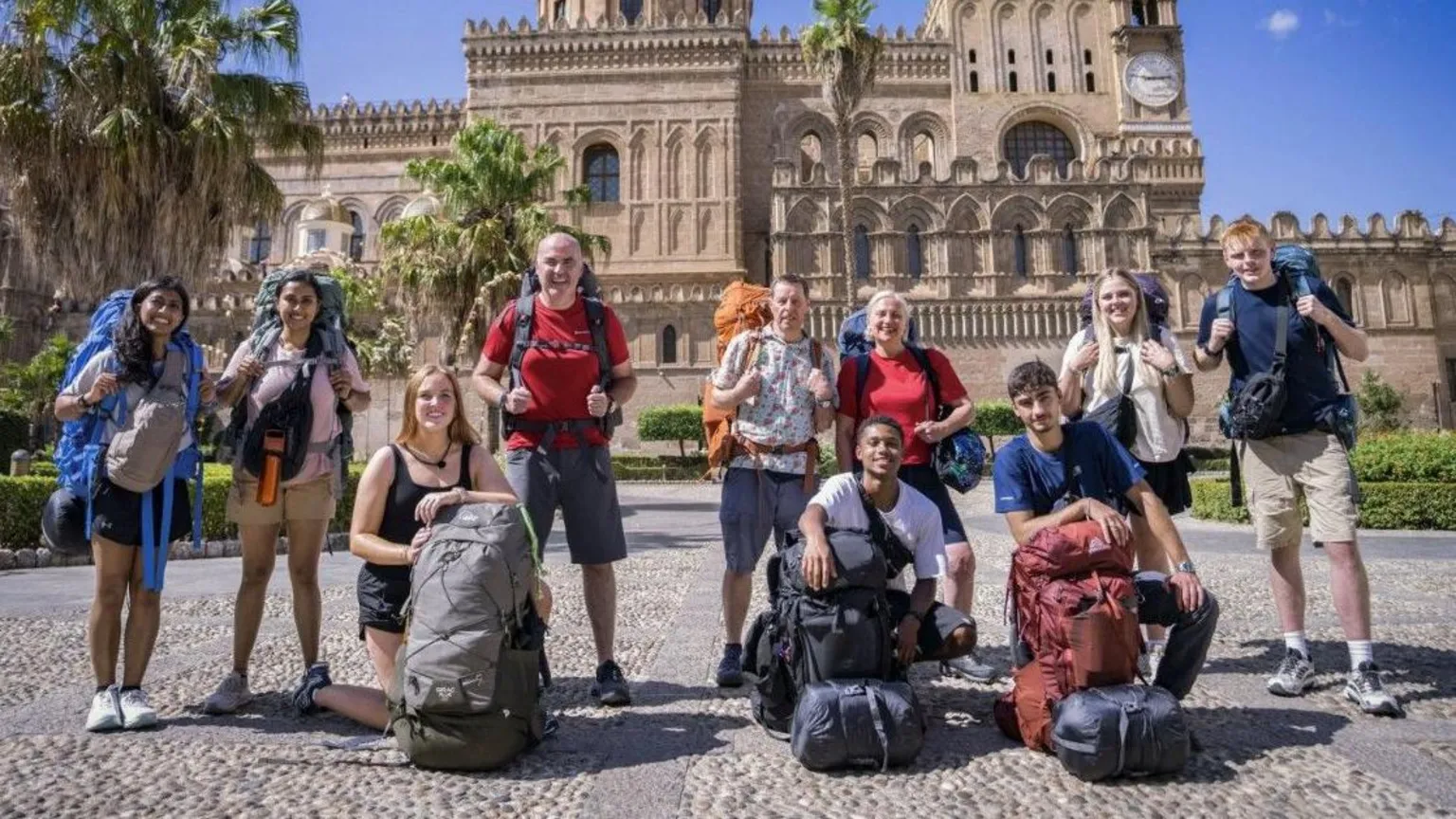 BBC/Studio Lambert The 10 contestants taking part in the latest series of Race Across the World are gathered for a photograph in front of palm trees and a Gothic-style building. They all have large backpacks and are smiling at the camera. 