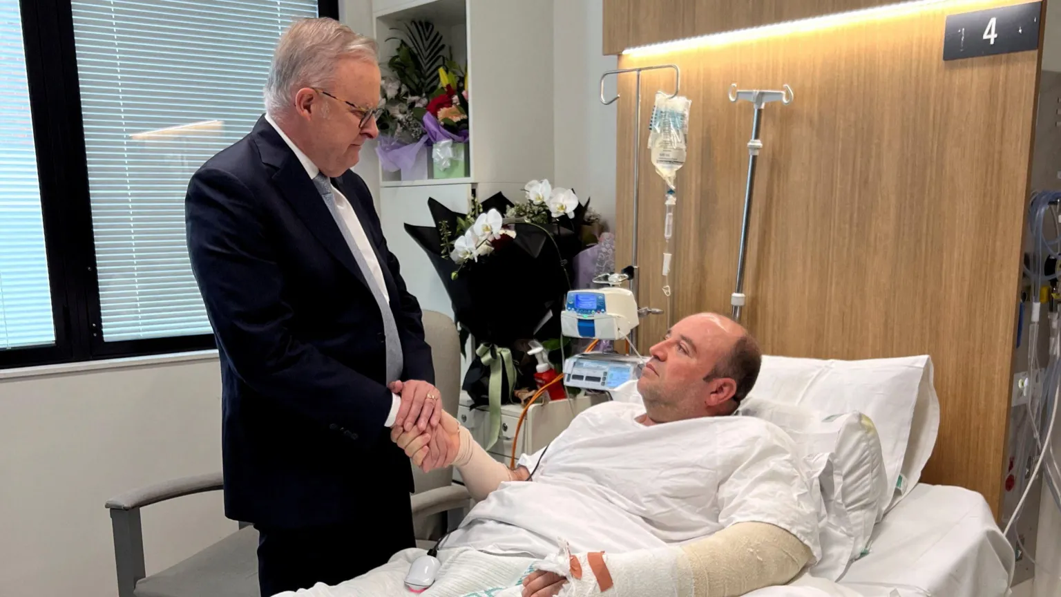  A man in a dark suit shakes the hand of a man lying in a hospital bed