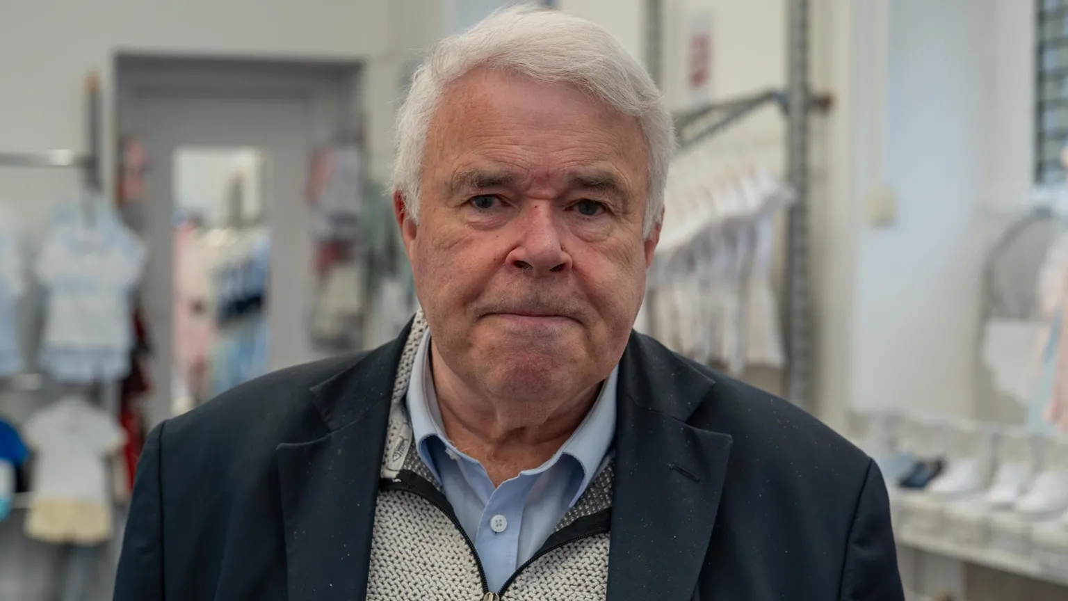 A man with grey hair wearing a blue shirt and suit jacket. He is standing in a shop and items of baby clothing are visible behind him.