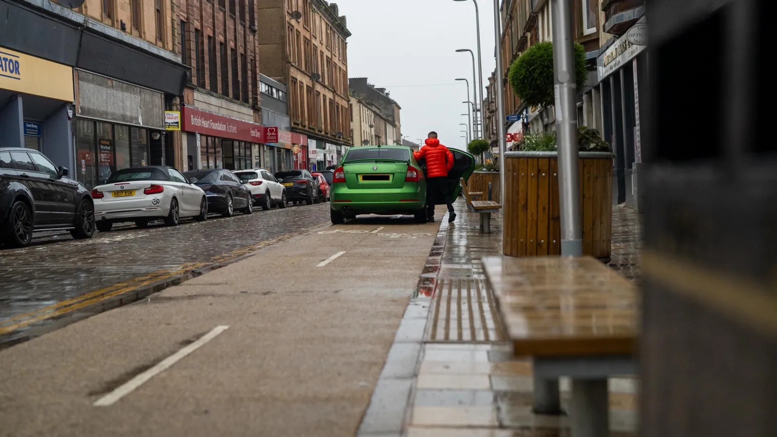 A green car parked on a two way cycle lane with a person wearing an orange jacket getting in to it.