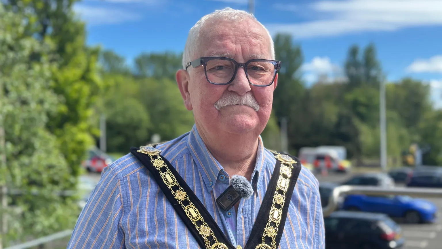Oliver McMullan has white hair and a white moustache and glasses. He is wearing a blue shirt with think salmon stripes and over the top he is wearing his mayoral chain of office. He is standing in a car park, trees can be seen out of focus behind him.