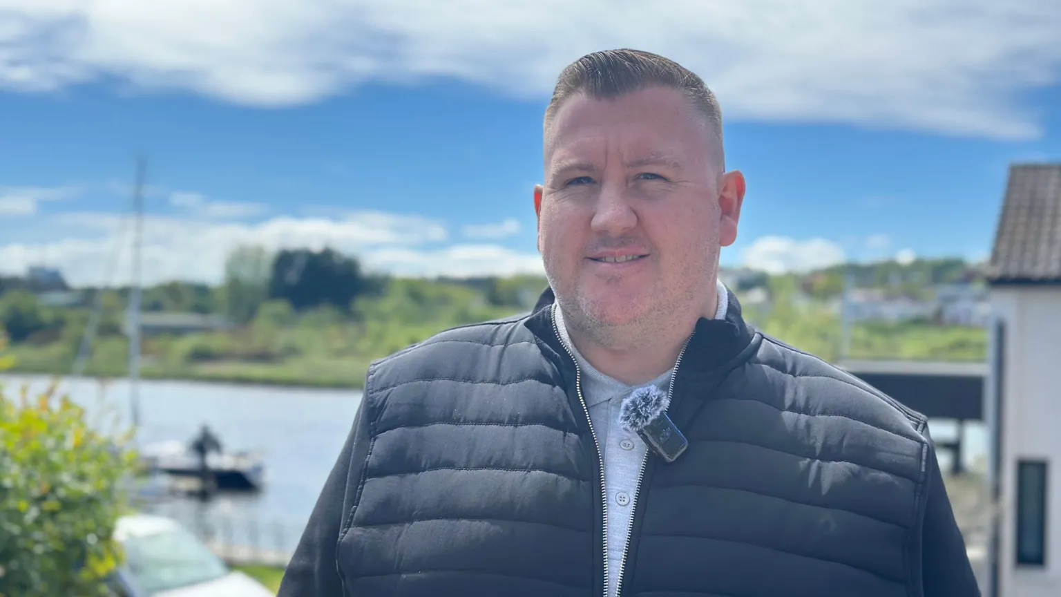 Eoin Henney is smiling at camera - he has short brown hair and is wearing a black jacket and grey polo shirt. He is standing outside and a river can be seen behind him with a sailboat.