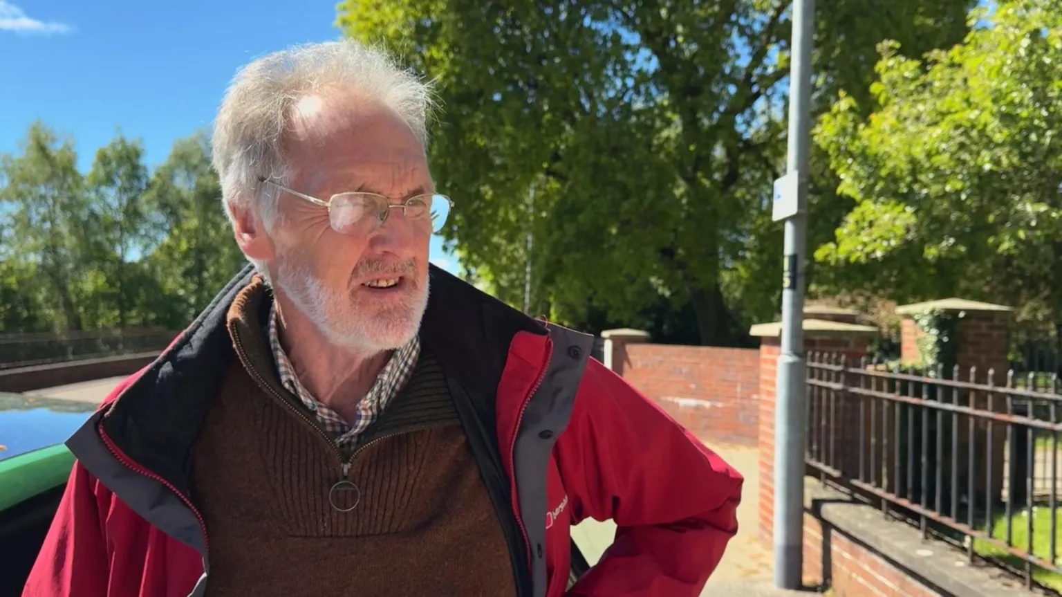 Roger has frizzy white hair and a short white beard and is wearing a red coat, brown jumper and chequed shirt. He is standing at the side of the street beside a red wall with a rusted iron fence. There are trees in the background.