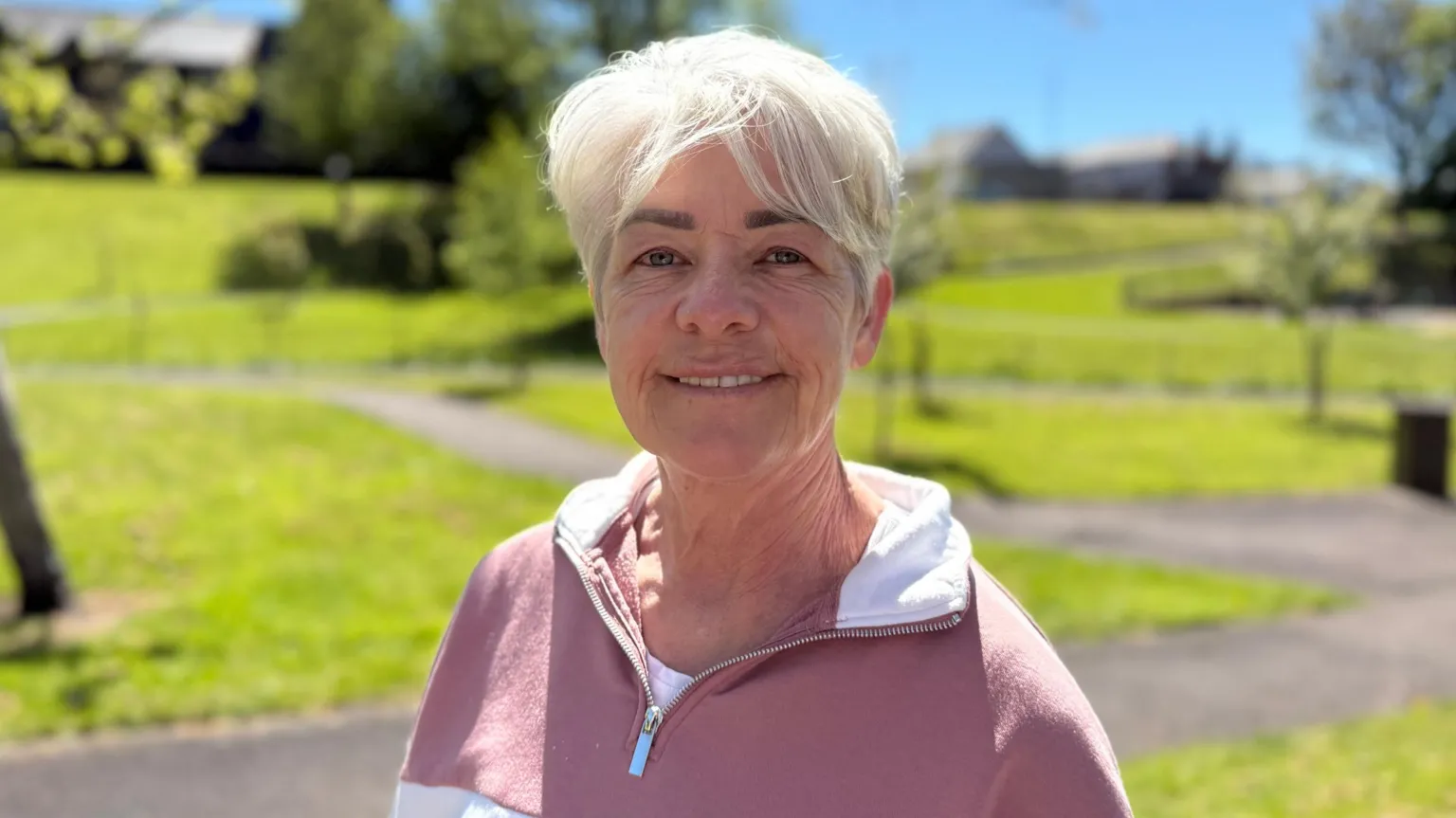 Juliete has white hair and is smiling at camera wearing a light pink and white quarter zip jumper sweater. She is standing in a park, grass and tarmacked paths can be seen behind her.