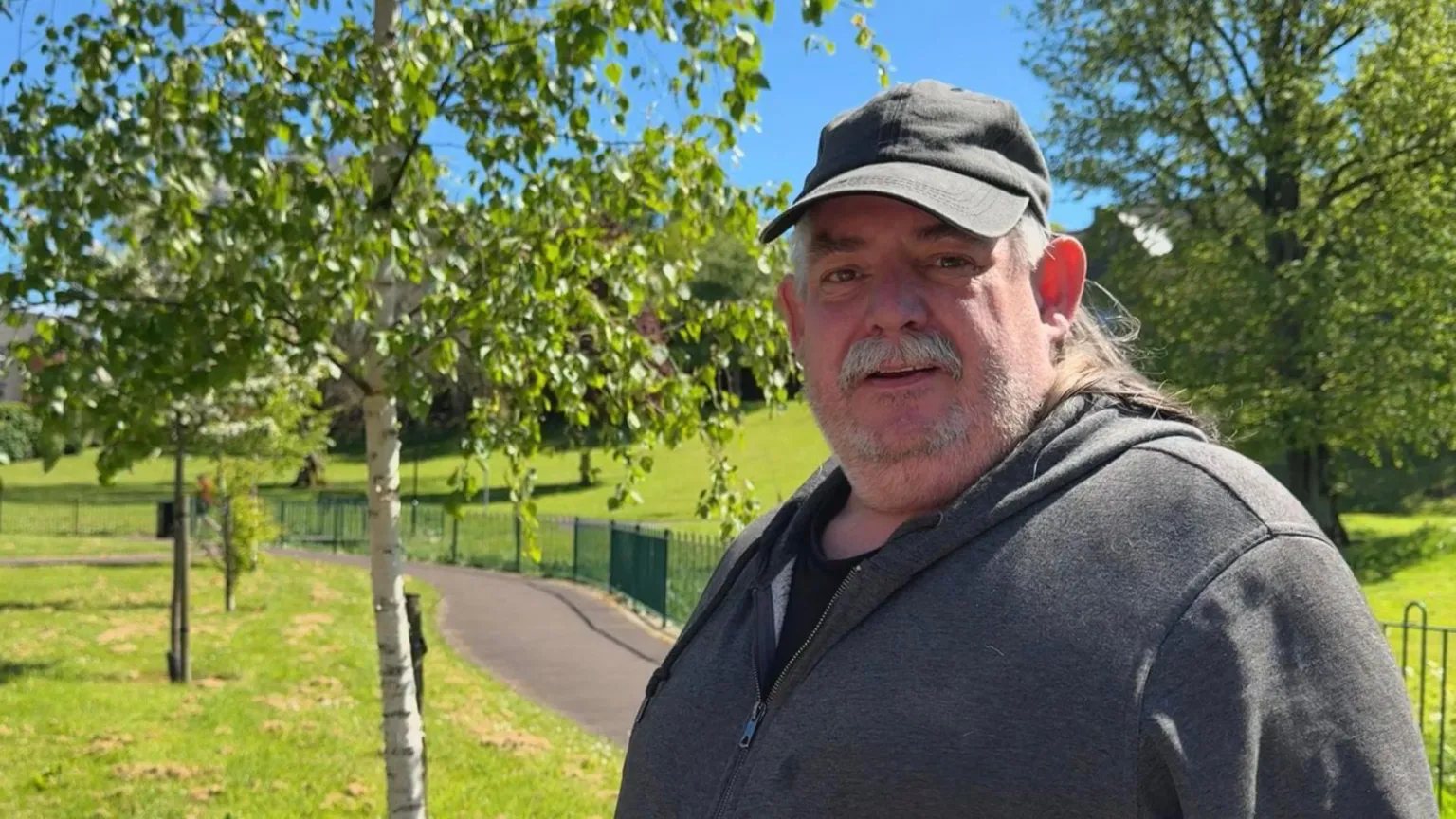 Tony has a grey beard. He is wearing a black baseball cap and a grey zipped jacket. He is standing in a park area with trees, grass, a footpath and green fencing.