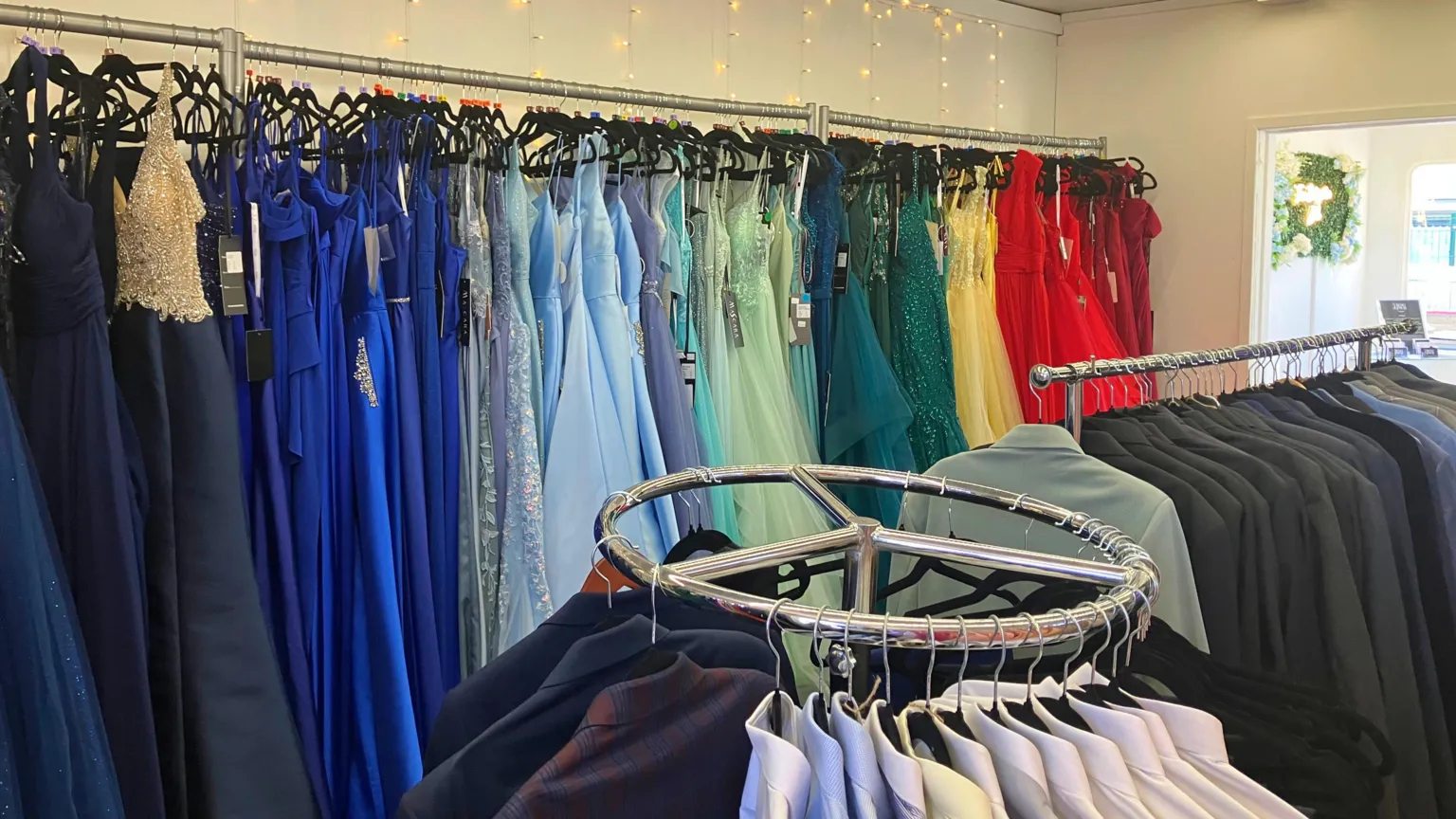 BBC/ Joanita Musisi A formalwear boutique with racks of dresses arranged in a colour gradient from dark blues to bright reds, alongside a circular rack of men’s suits and shirts, all set against a backdrop of warm string lights.