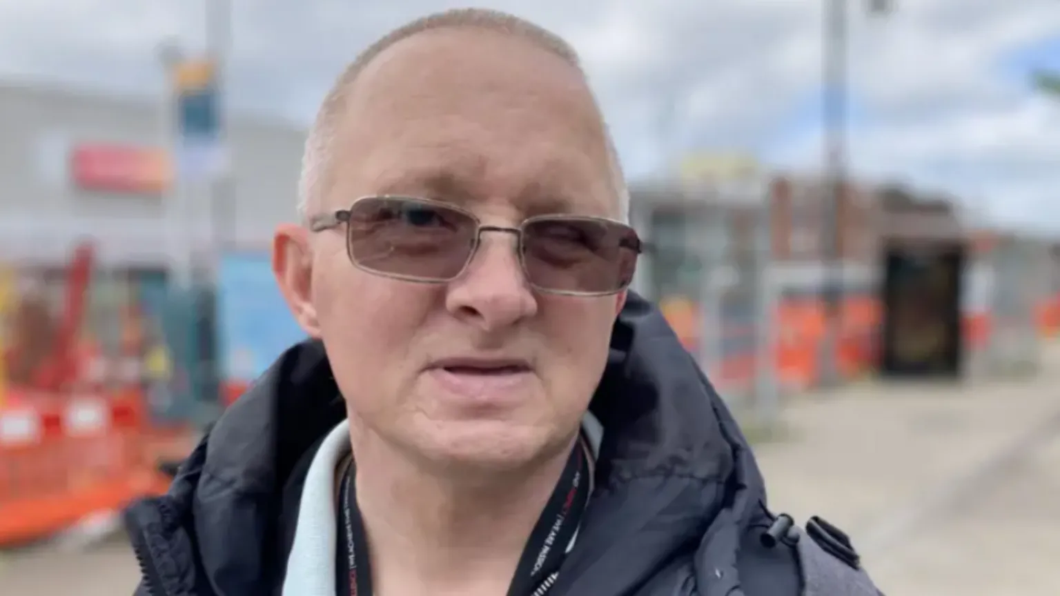 A man wearing tinted glasses looking into the camera. He is stood outside.