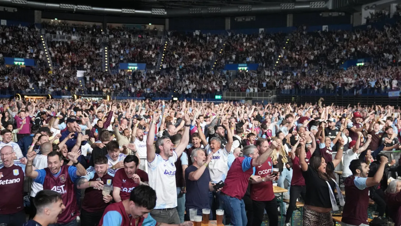  Claret and blue clad fans jump for joy in the arena. Most are in Villa tops. Many have their hands in the air at ground levels and in surrounding seating.