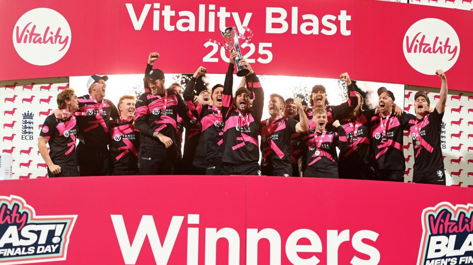 Lewis Gregory of Somerset lifts the Vitality Blast Men's Trophy, surrounded by cheering teammates, all of which are behind a red 'Winners' board.