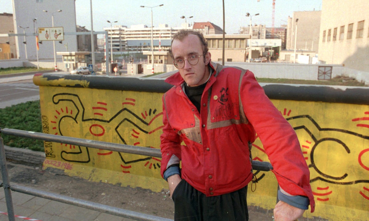 Keith Haring stands in front of a section of the Berlin Wall he painted with one of his signature crawling babies in Berlin, 1986.