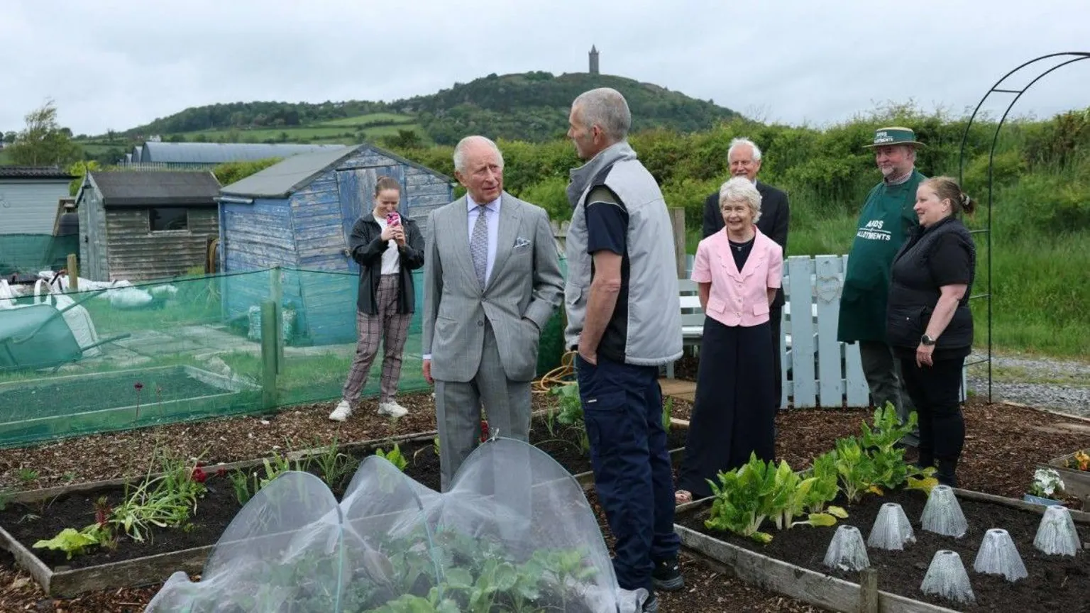  King Charles III standing in an allotment. There is a group of men and women standing behind him and he is talking to a man. 