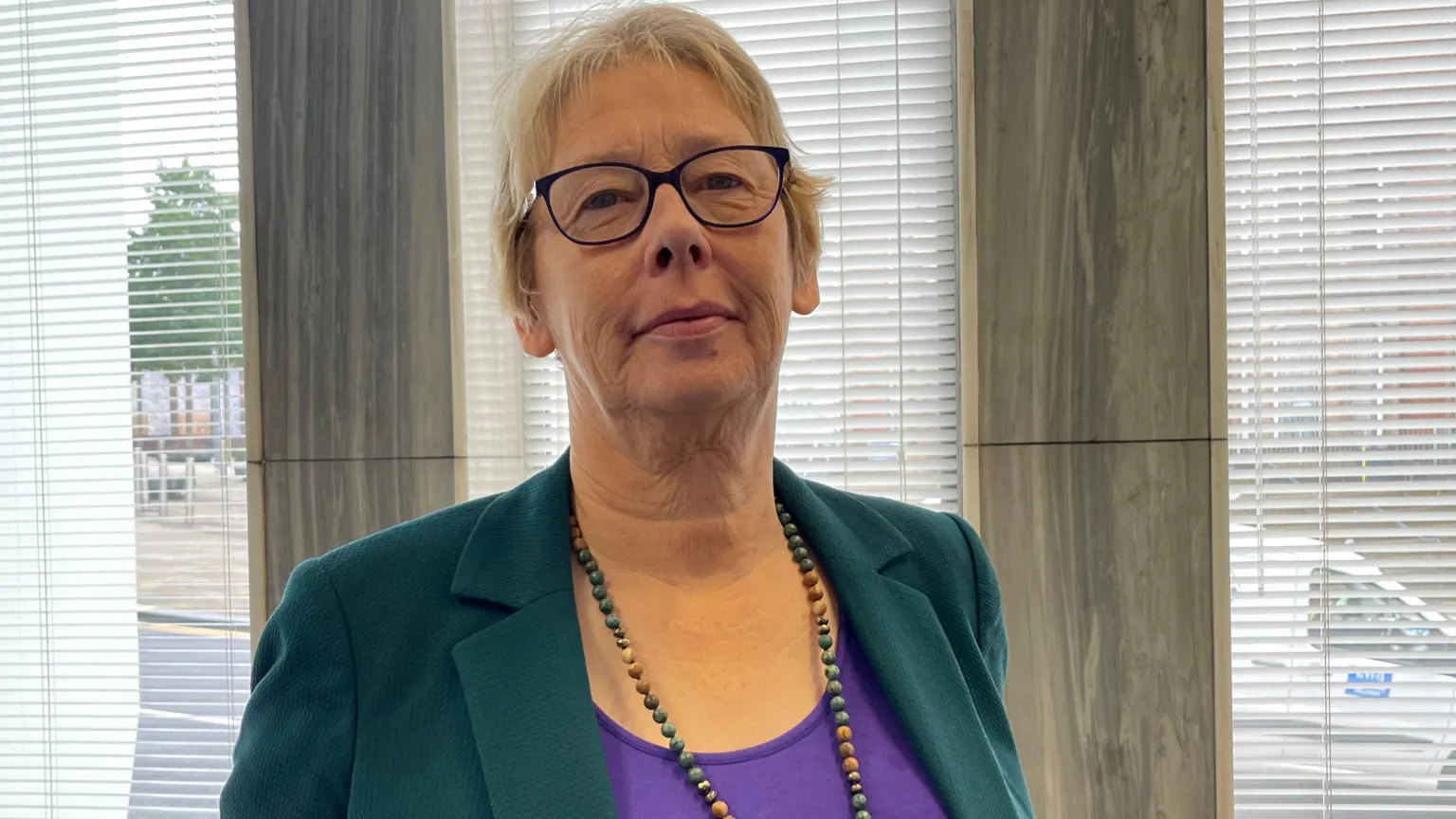 Jane Ashworth - a woman with short blonde hair, glasses, a beaded necklace, purple top and green jacket - stands in front of a window.