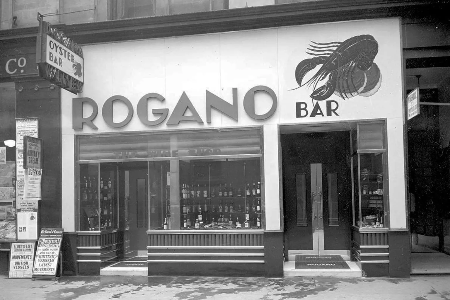 Glasgow Museums and Libraries Collections Period black and white photograph of the exterior of Rogano oyster bar in Glasgow, showing the ROGANO BAR signage, a painted lobster graphic, and window displays filled with bottles and glassware.