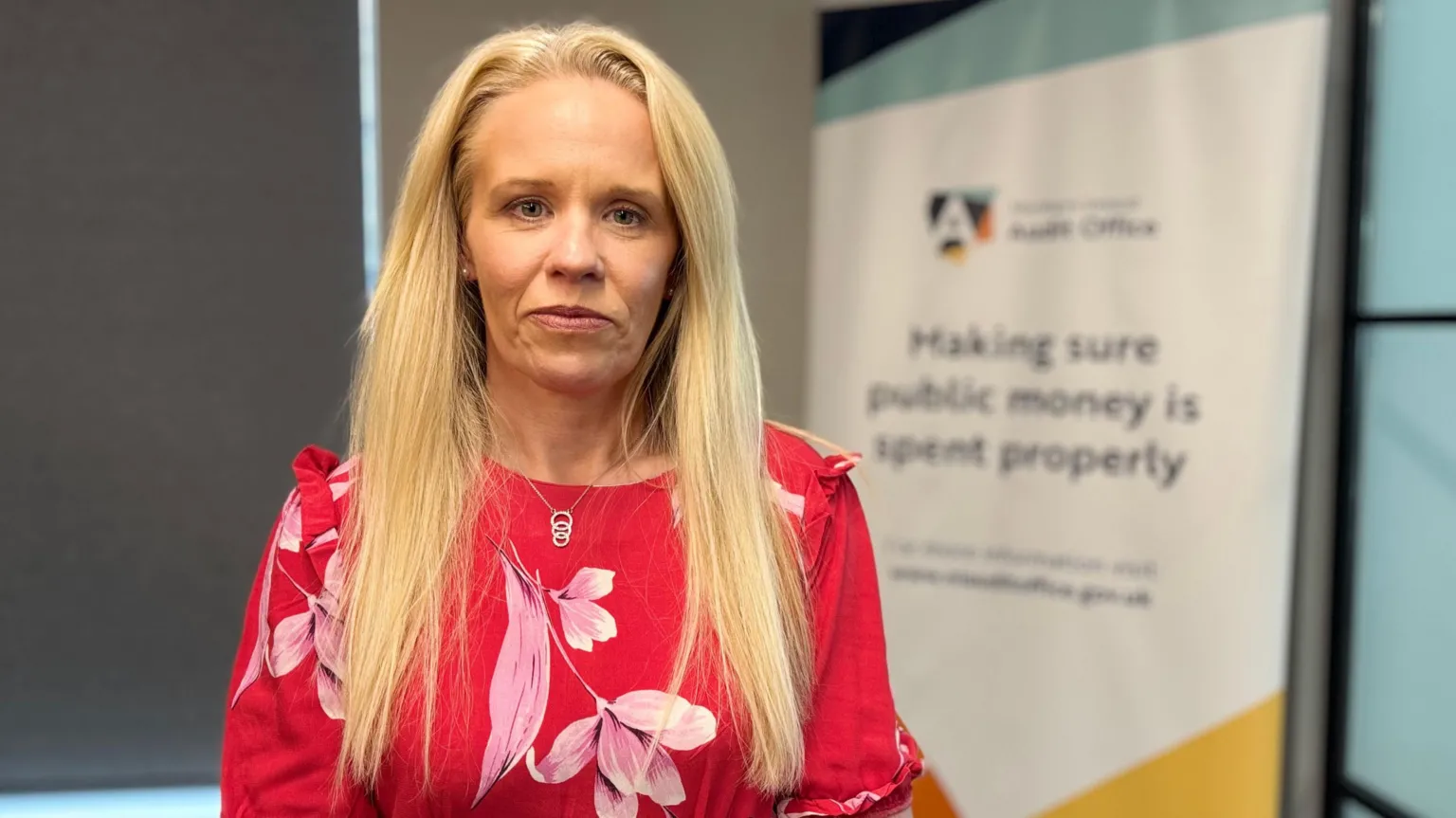 Dorinnia Carville is wearing a red top with pink lilies over it. Her long hair is blond. A board is out of focus behind her. 