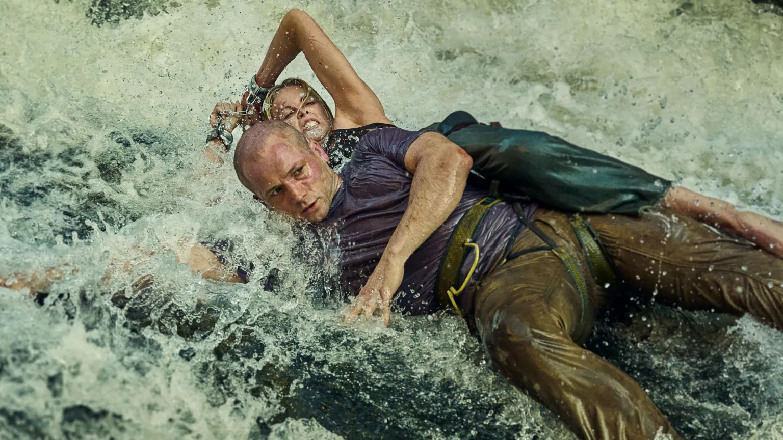 Netflix Egerton and Theron both cling to a large boulder in a river, with water crashing over them. Egerton wears a blue t-shirt and brown trousers, while Theron wears green trousers