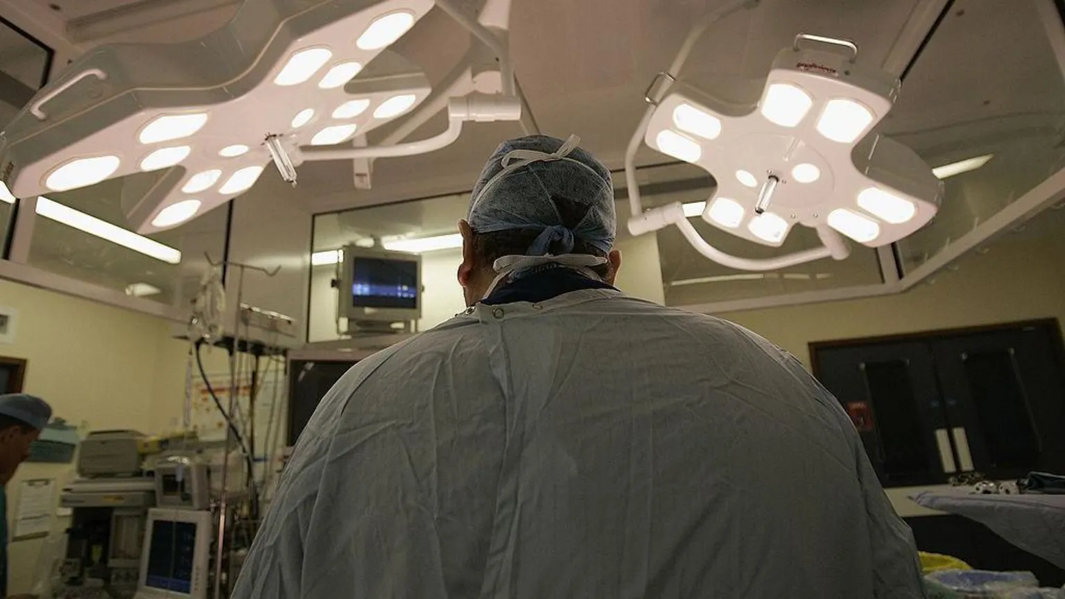 Surgeon seen from the back in operating theatre under bright overhead lights in a surgical gown