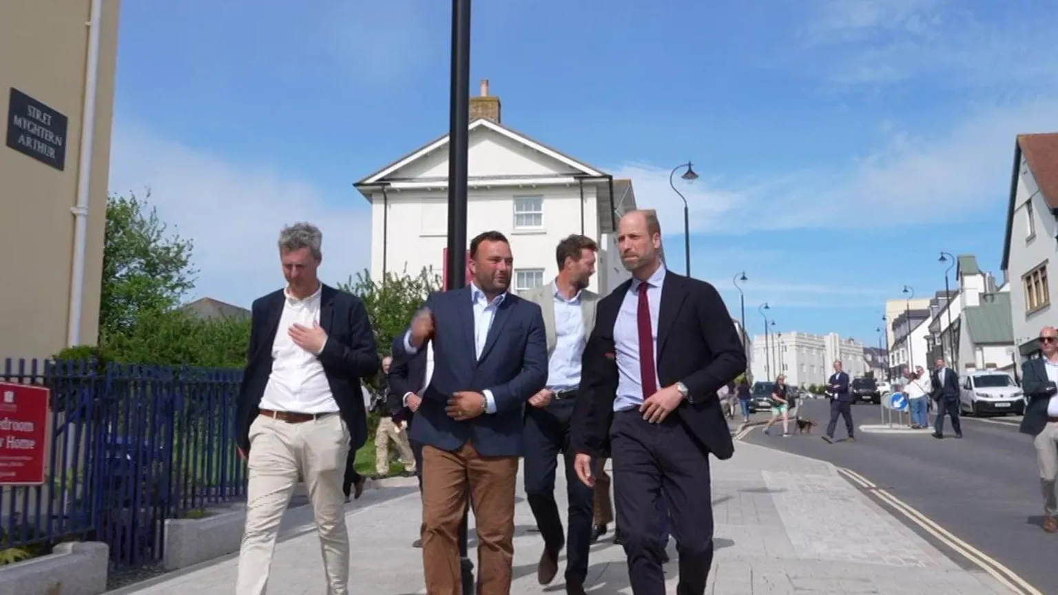 Prince William walking with two men along the street. He is talking to them and walking at the same time. There is a road to his right, and houses behind him and to right.