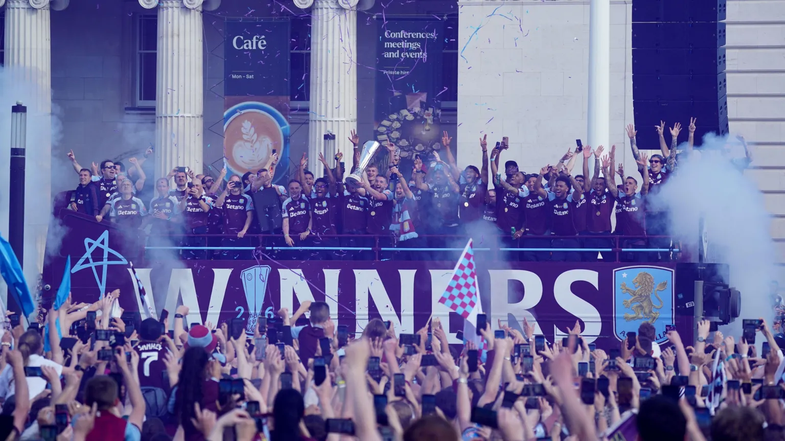 Villa players on a bus lift the trophy as fans raise their arms below.
