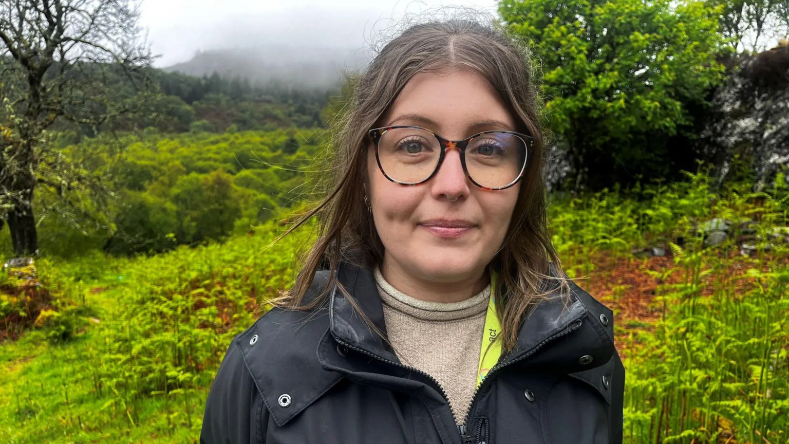 Heather Morrison is wearing large glasses, and a brown top with a black waterproof jacket. She is standing among grasses and ferns in a forest