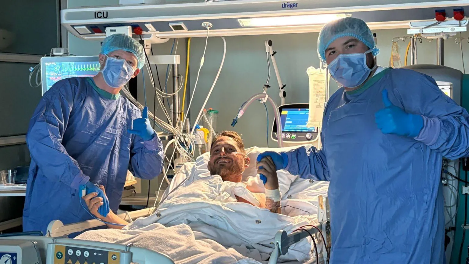 Rhys Thomas Rhys laying in a hospital bed with tubes coming out of him leading to machines. Two adult men stand either side of the bed holding his hand. They are wearing blue medical protective clothing.