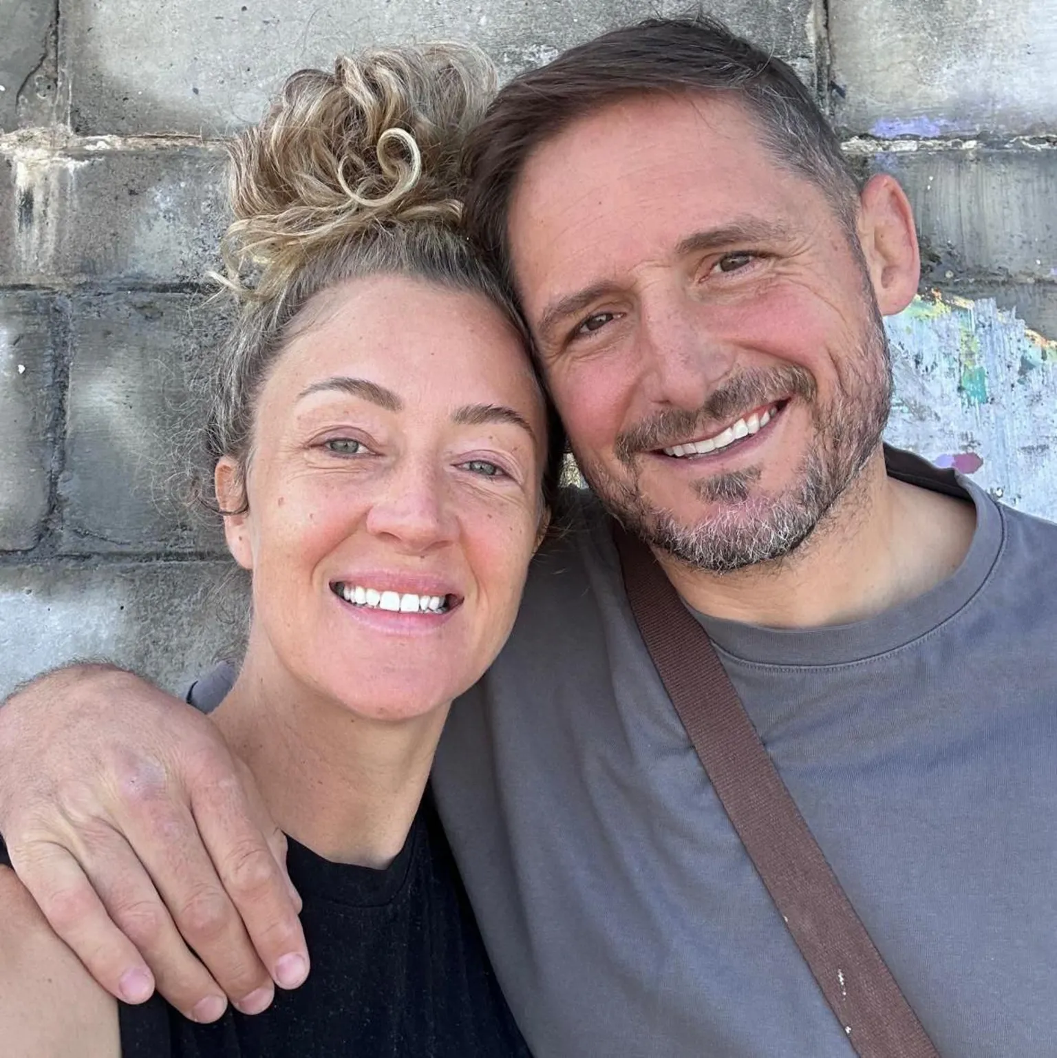 Rhys Thomas Selfie of Thomas and Kez stood in front of a grey brick wall. Kez wears a black top and has blonde curly hair. Thomas wears a grey t-shirt and a brown leather bag carrying his left ventricular assist device.