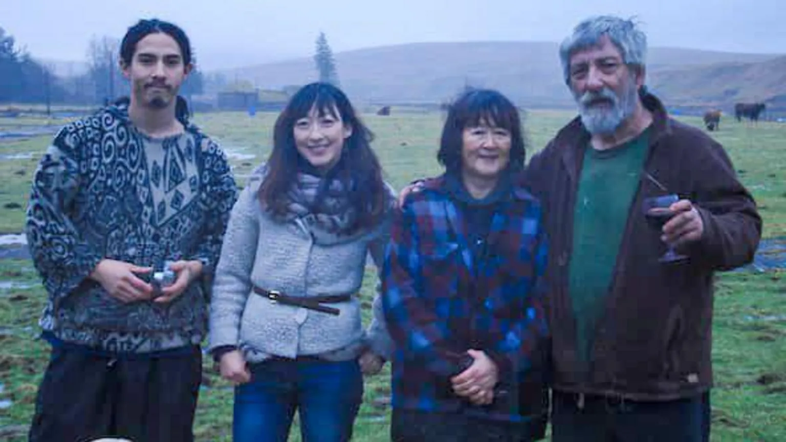 Holmes Family A family picture with, from left to right, a young man with long, dark hair pulled back in a patterned jumper, a woman with long, dark hair in a grey jacket with a black belt around her waist, a woman with shorter dark hair and a blue and black coat and a man with grey hair and a beard in a dark jacket holding a drink.