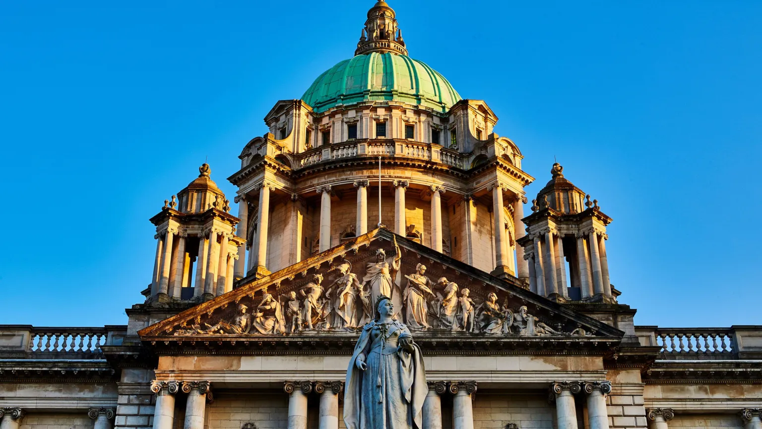  A close up of Belfast City Hall with a blue sky. A statue of Queen Victoria is in the centre. The building has numerous pillars and a green dome. 