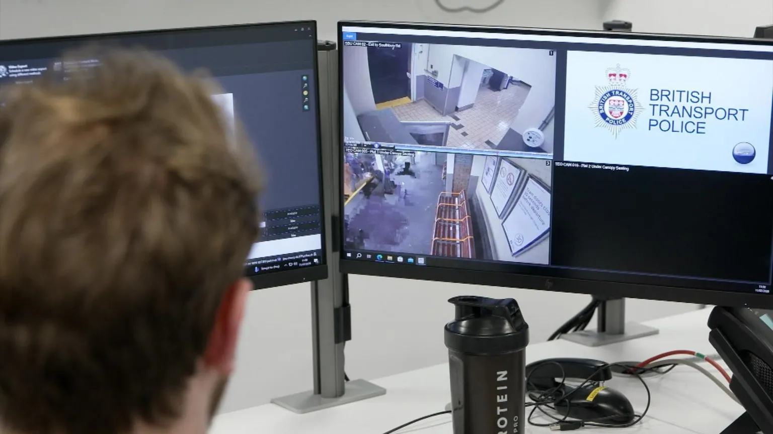 A BTP officer looks at two computer monitors. The one on the right has two CCTV images on it as well as the BTP logo
