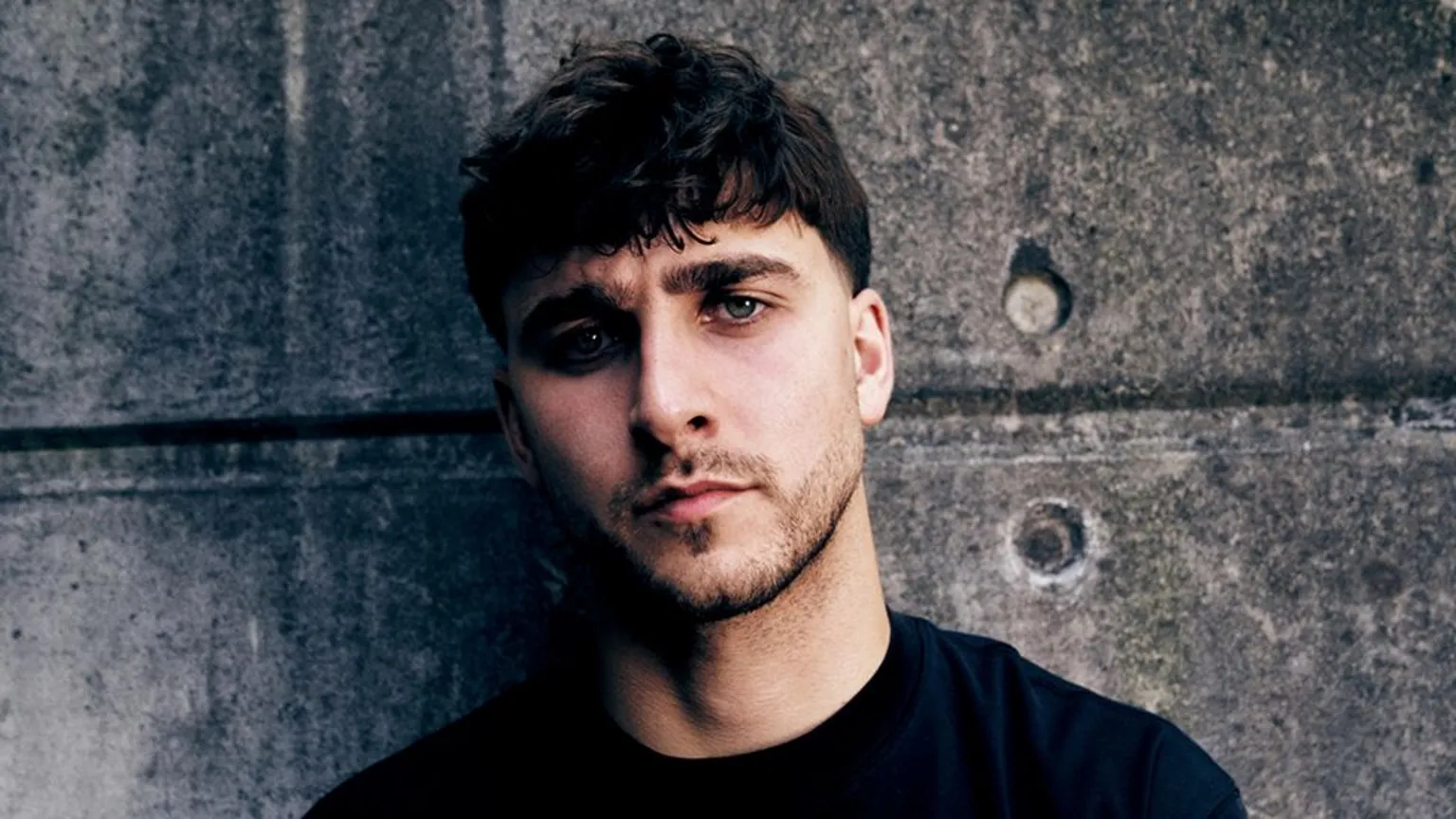 Joe Magowan A man wearing a dark coloured top, standing in front of faded grey concrete bricks.