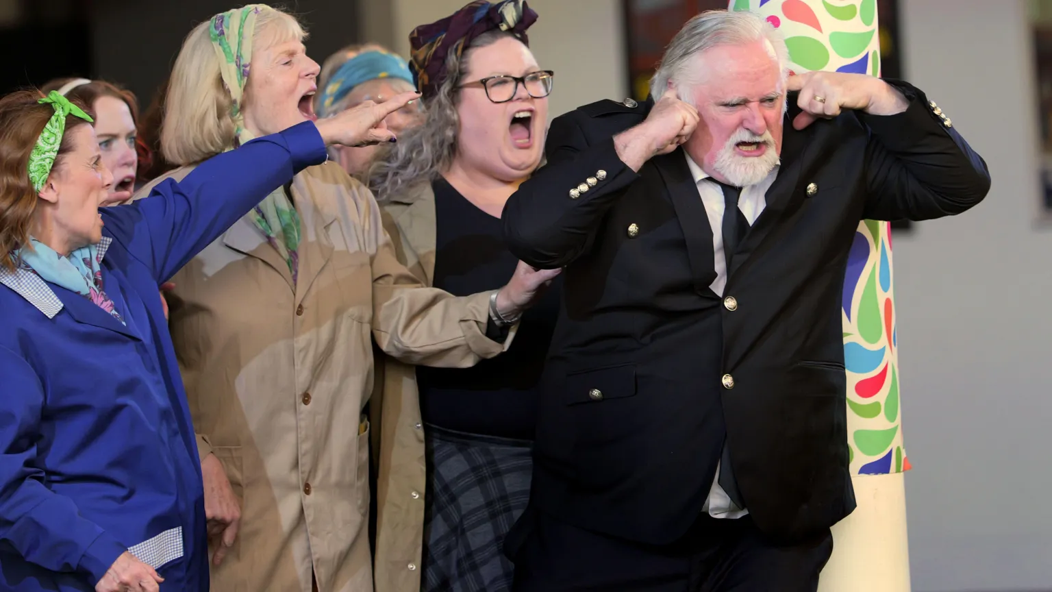 Sartorial Pictures A scene from Scottish Opera's production of Carmen, with several women singing/shouting and pointing at a man in a suit, who has his fingers in his ears as if blocking them out.
