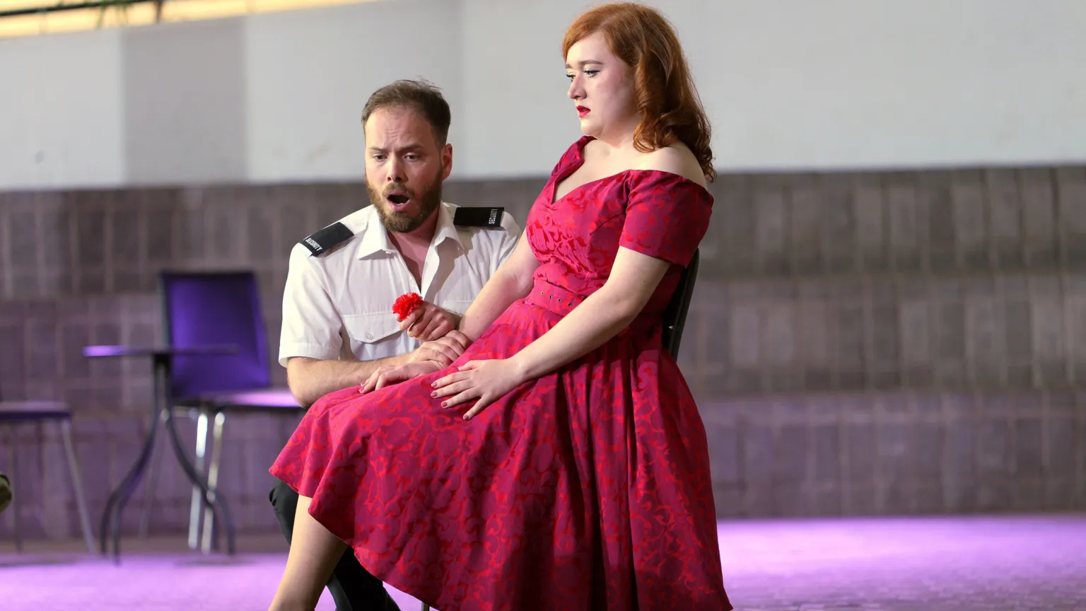 Sartorial Pictures Xavier Hetherington and Rosie Lavery on stage during Carmen. He is wearing a white shirt, has short dark hair and a beard and is kneeling next to Rosie while holding a rose. She is wearing a red dress and has red hair. 