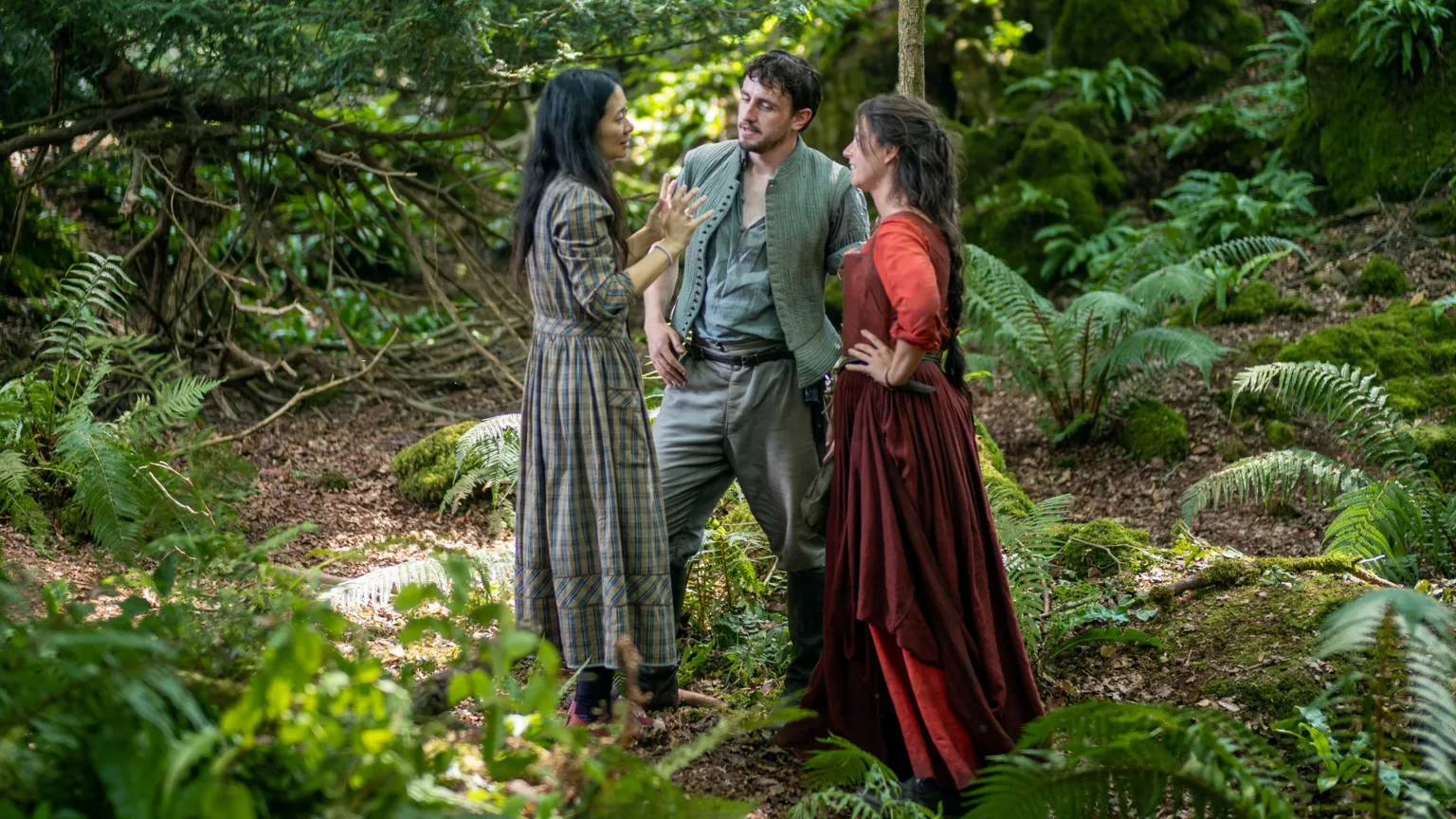 Universal Pictures [L-R] Behind-the-scenes image showing director Chloé Zhao on the set of Hamnet with Paul Mescal and Jessie Buckley They are standing in the middle of a lush green forest. Chloe with long black hair in long checked grey dress Paul in moss green top and trousers Jessie in dark red dress