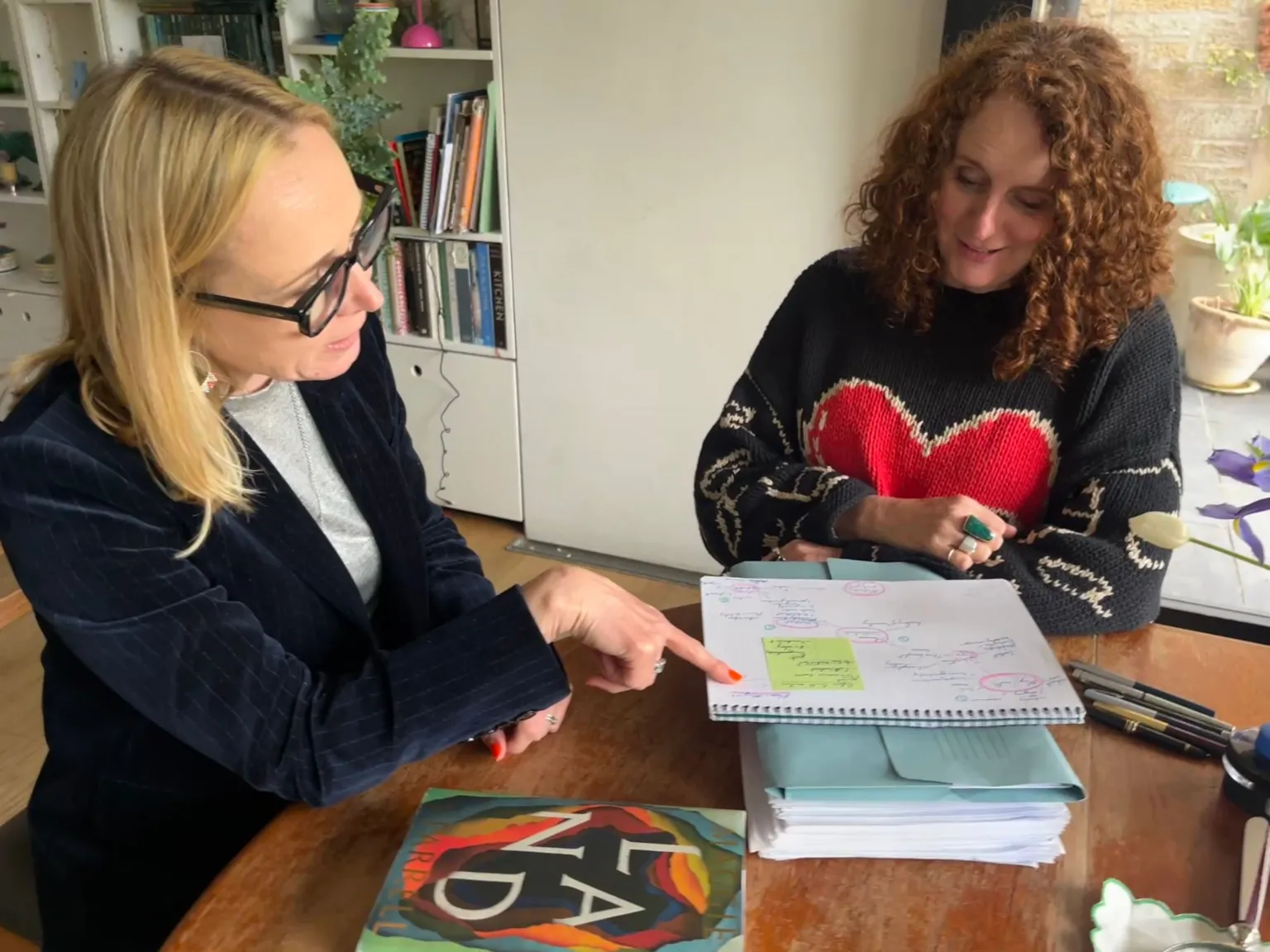BBC/Katie Razzall Katie Razzall on the left, blonde hair, glasses in dark blue velvet jacket seated at a table, pointing to note book. Maggie O'Farrell, auburn curly hair in dark jumper with red heart in the middle. Book shelves behind them on left Door to garden with plants in pots on the right