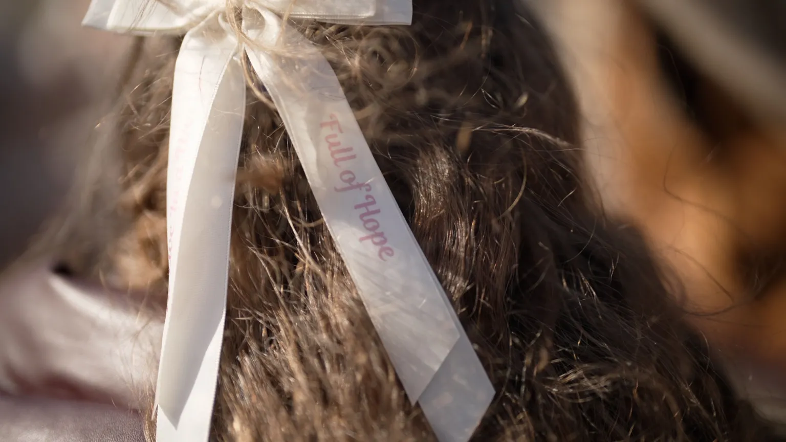 The back of a girl's head. She has brunette hair with a cream hair bow that has the words 'Full of hope' on it.