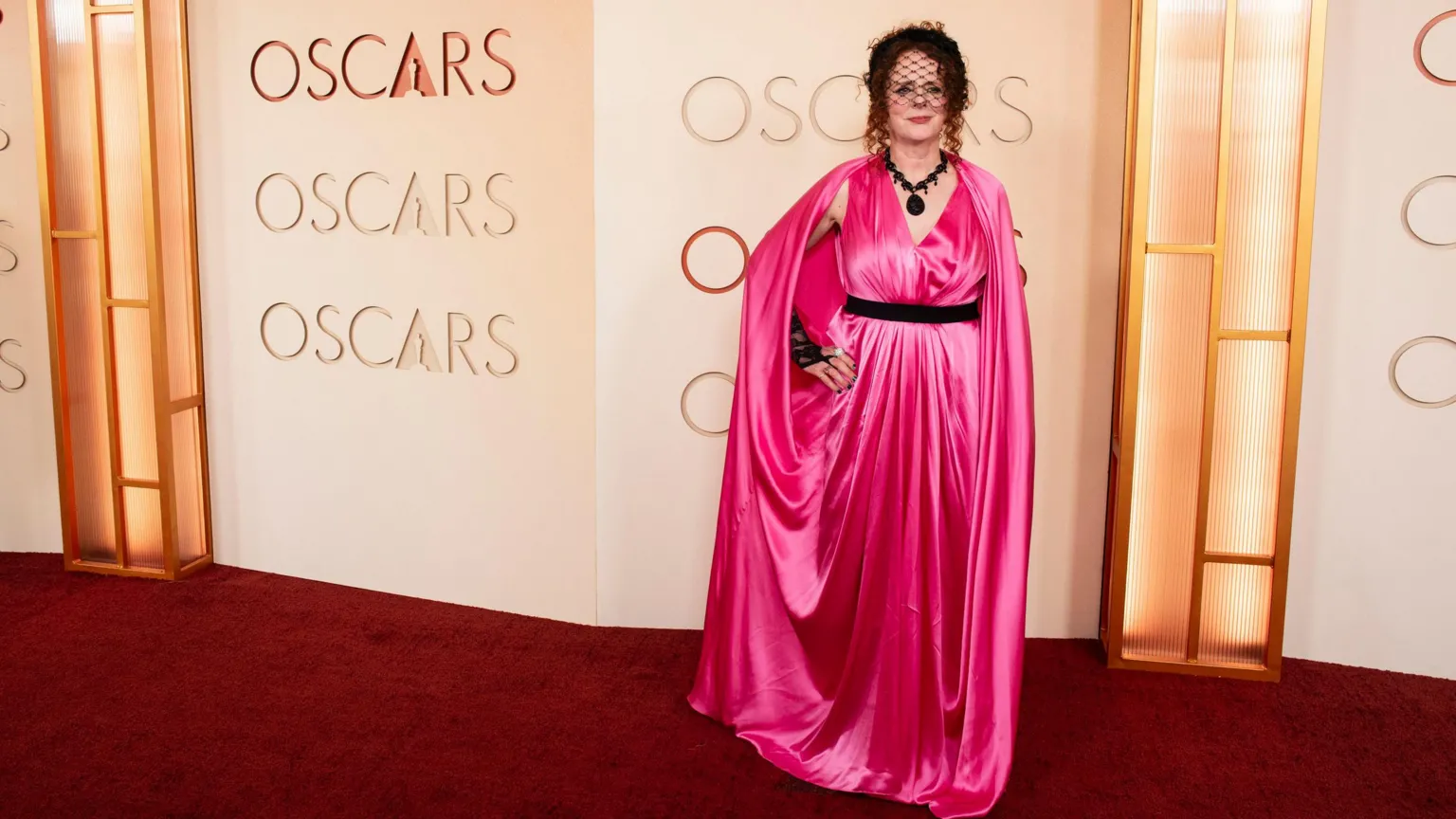 Maggie is bright pink long satin dress, black belt, black necklace & little veil hat. Posing behind the word Oscars.